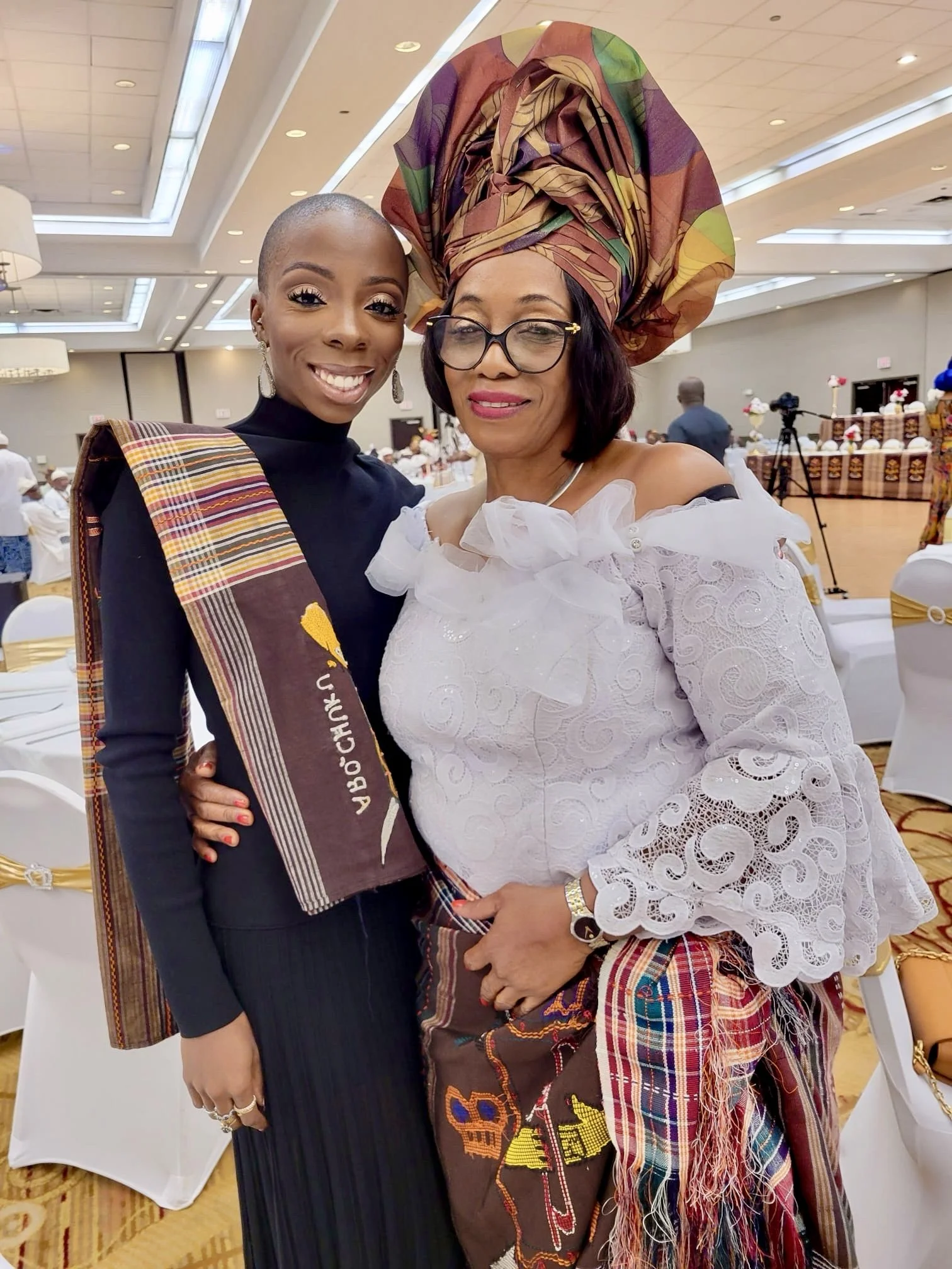 Two women in traditional African attire smiling at a celebration in a banquet hall. One woman wears a headwrap and glasses, and the other is dressed in a black outfit with a colorful sash.