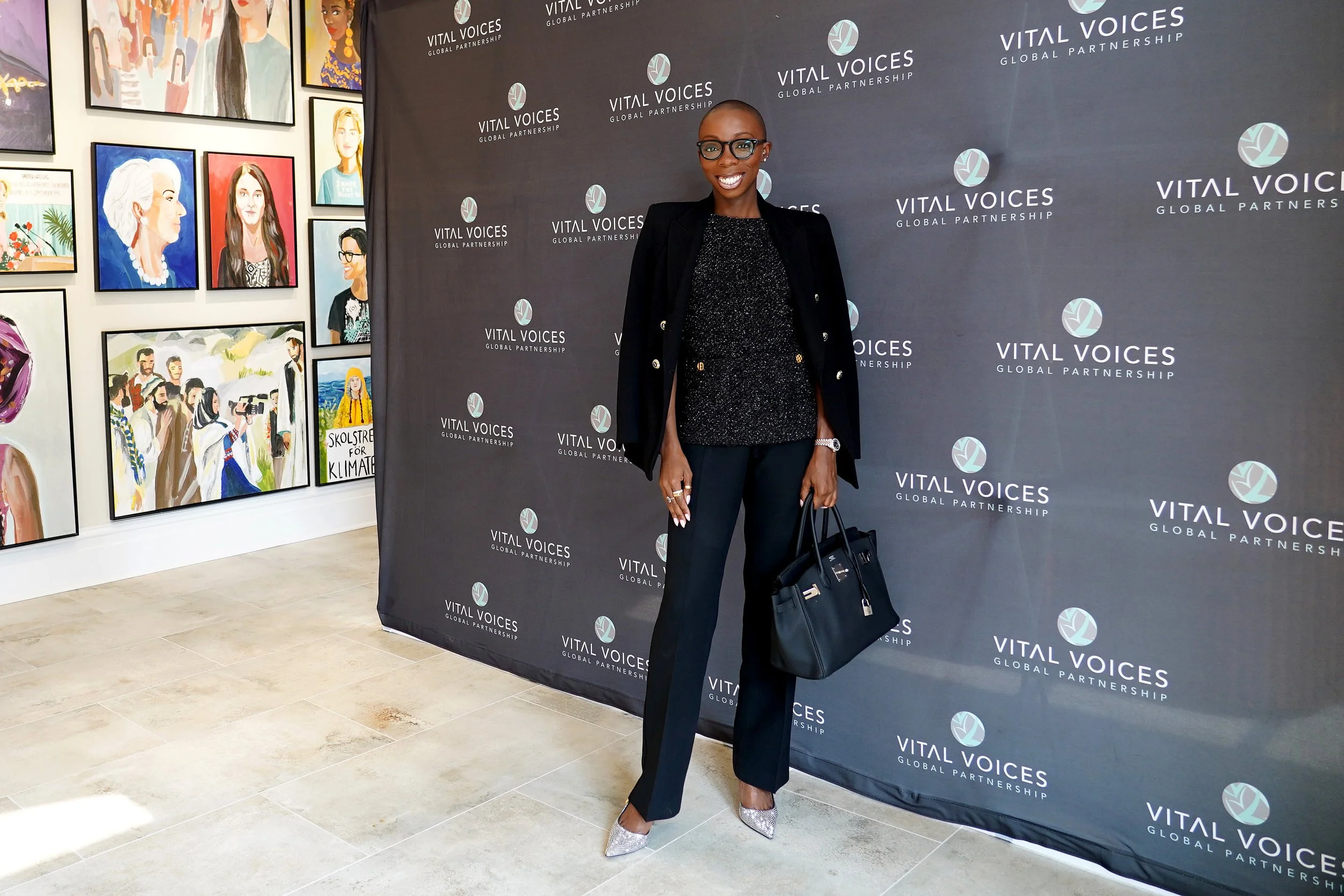 A woman dressed in black, wearing glasses, and holding a dark handbag, standing in front of a backdrop that reads 'Vital Voices Global Partnership'. The backdrop features a repeated logo pattern. The woman is smiling and posing for the camera in an a