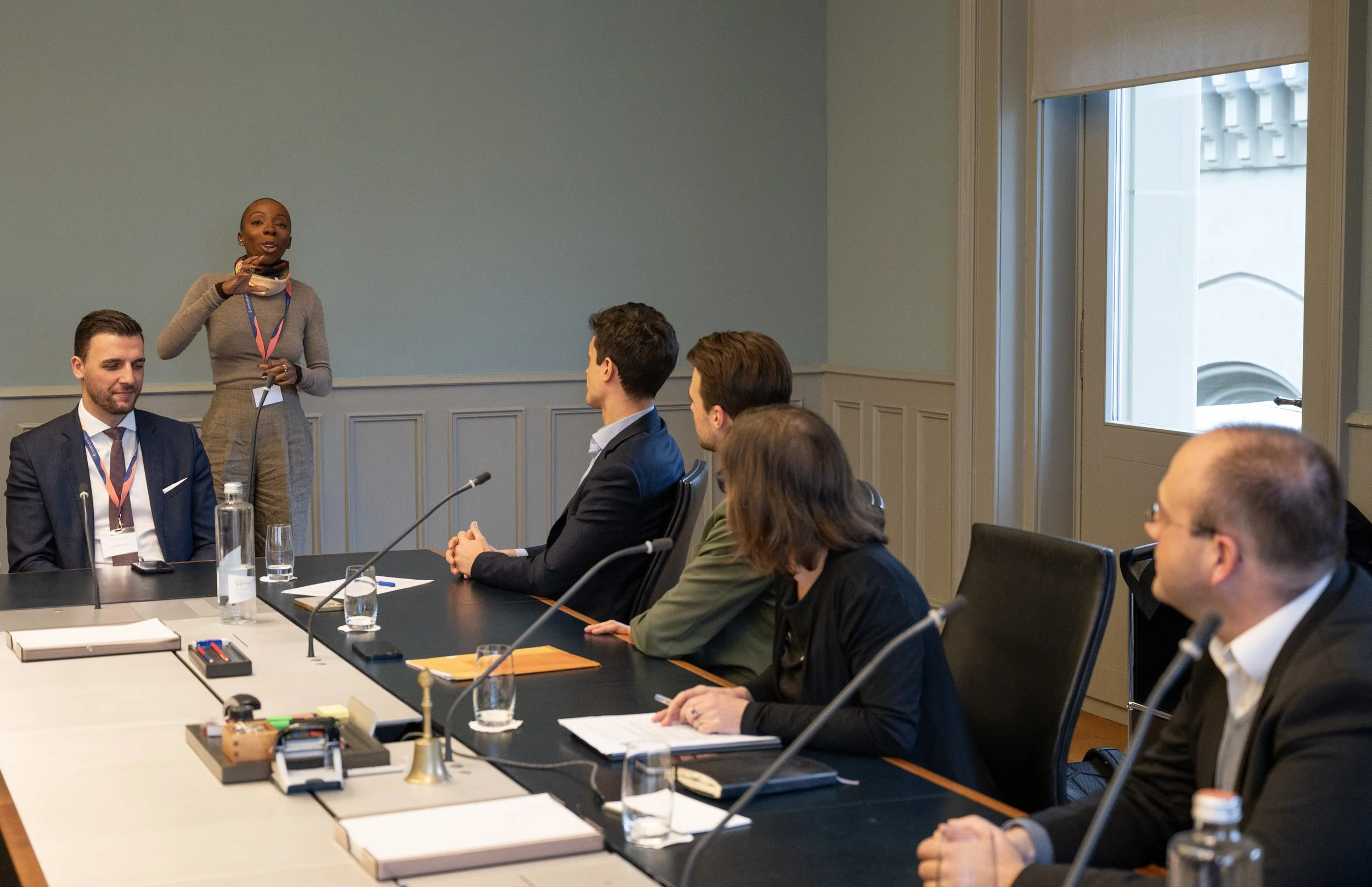 A woman in a gray outfit is speaking to a group of five people seated at a conference table in a meeting room with light blue walls and large windows.