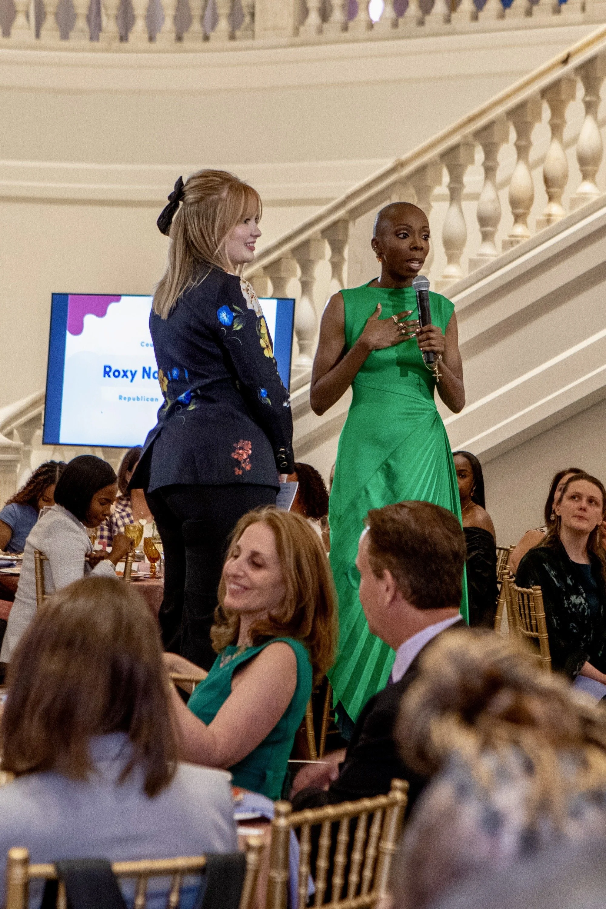 Two women speaking on stage at a formal event, one in a green dress holding a microphone, and the other in a dark floral blazer standing beside her. Audience members seated at tables listen attentively.