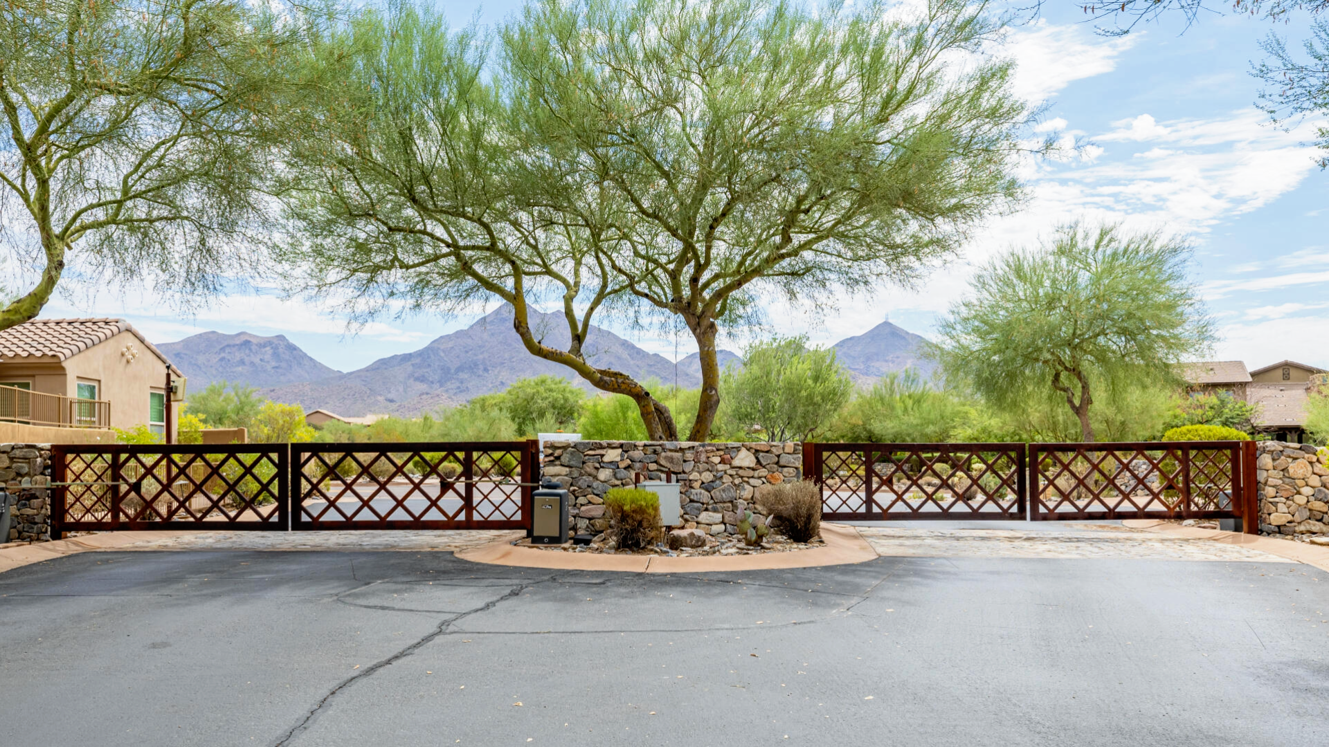 Residential neighborhood with a stone and wooden fence, desert trees, and mountains in the background under partly cloudy sky.