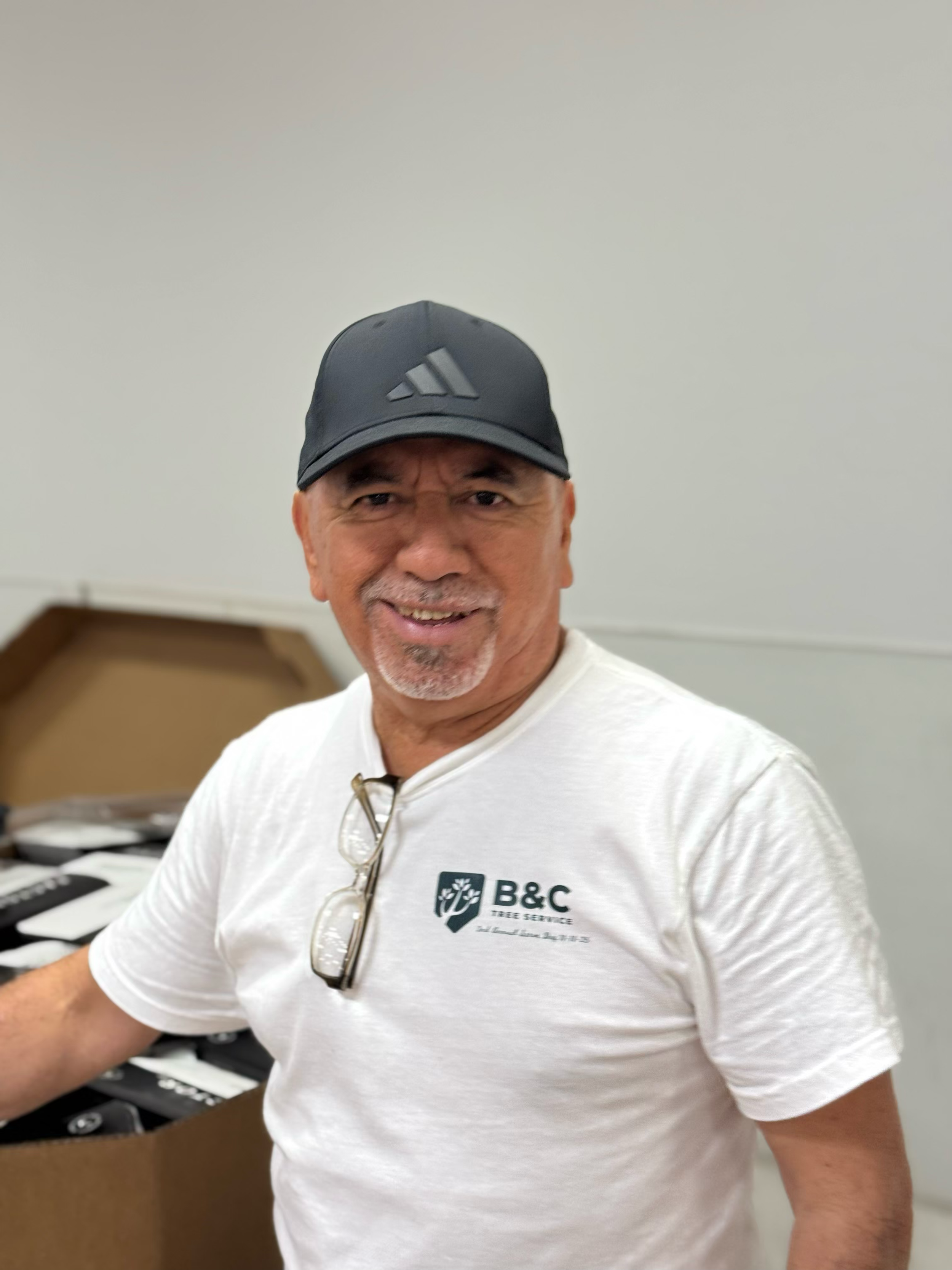 A smiling man wearing a black cap and a white t-shirt with a B&C Tree Service logo standing near a table or counter.