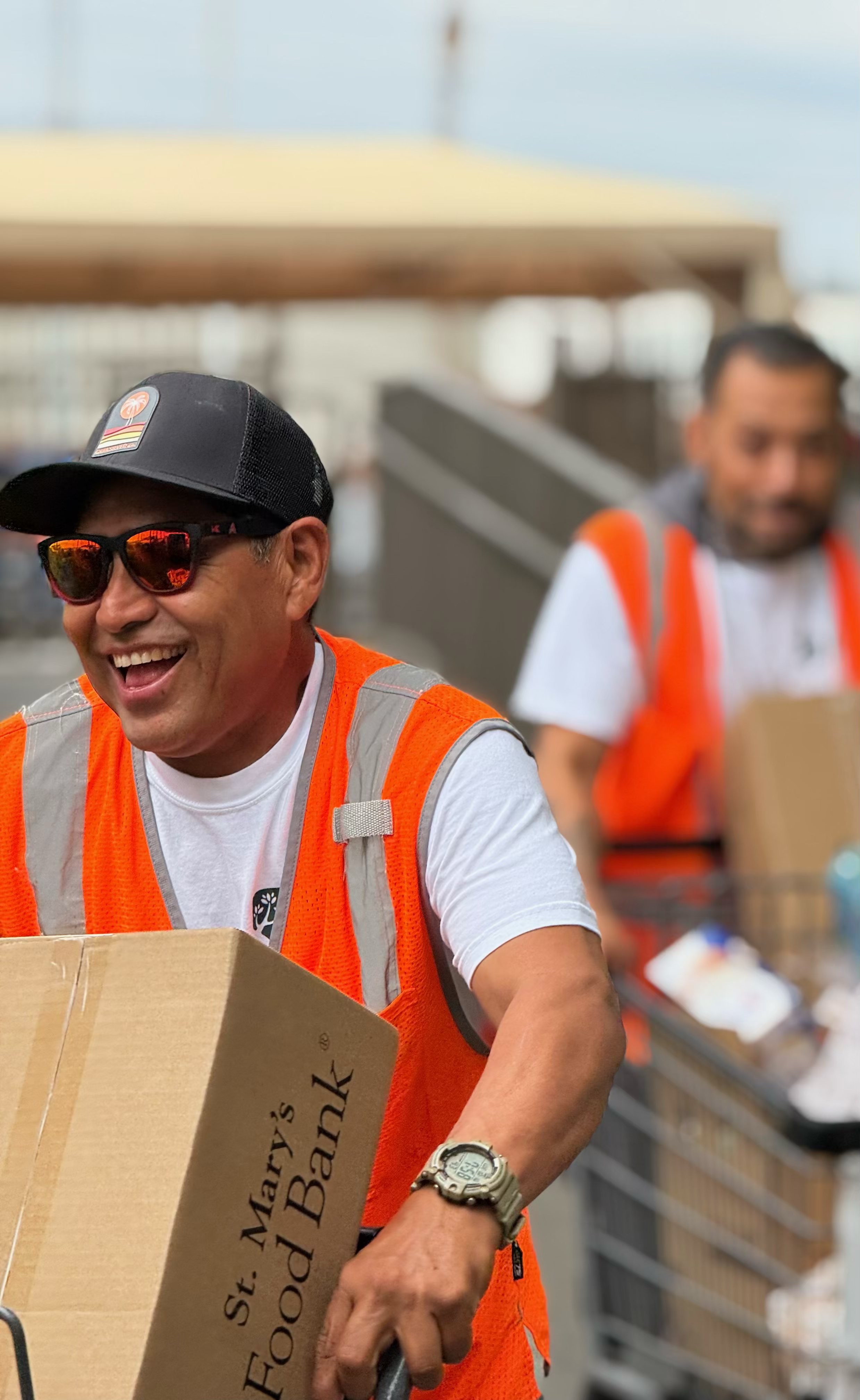 Two smiling men in orange safety vests working at a food bank or donation center, one holding a cardboard box, with food items in carts behind them.