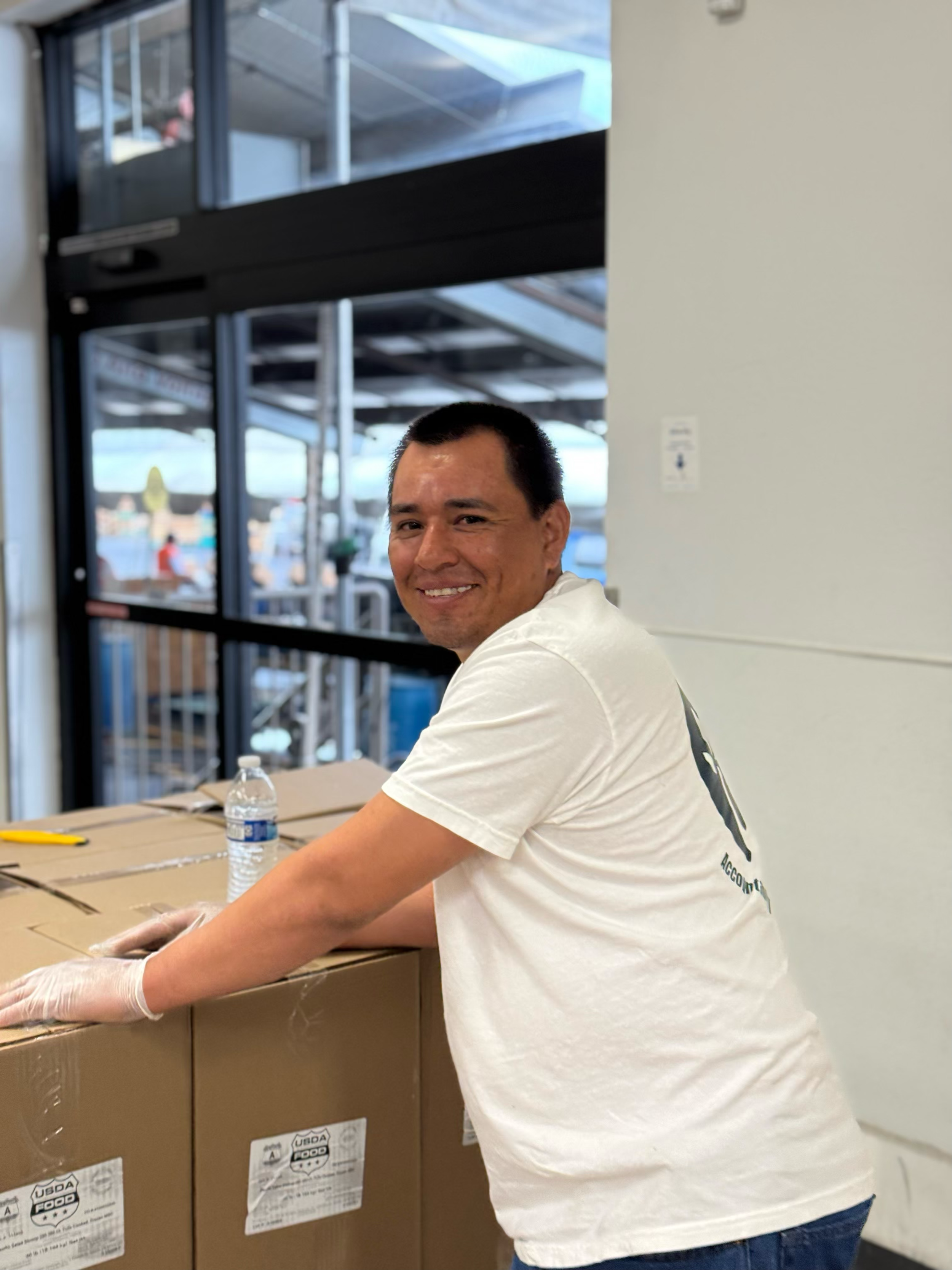A man wearing a white T-shirt and gloves is smiling and leaning on a cardboard box inside a store. There is a water bottle on the box, and the background shows a doorway with metal framing, outside view with people and equipment.