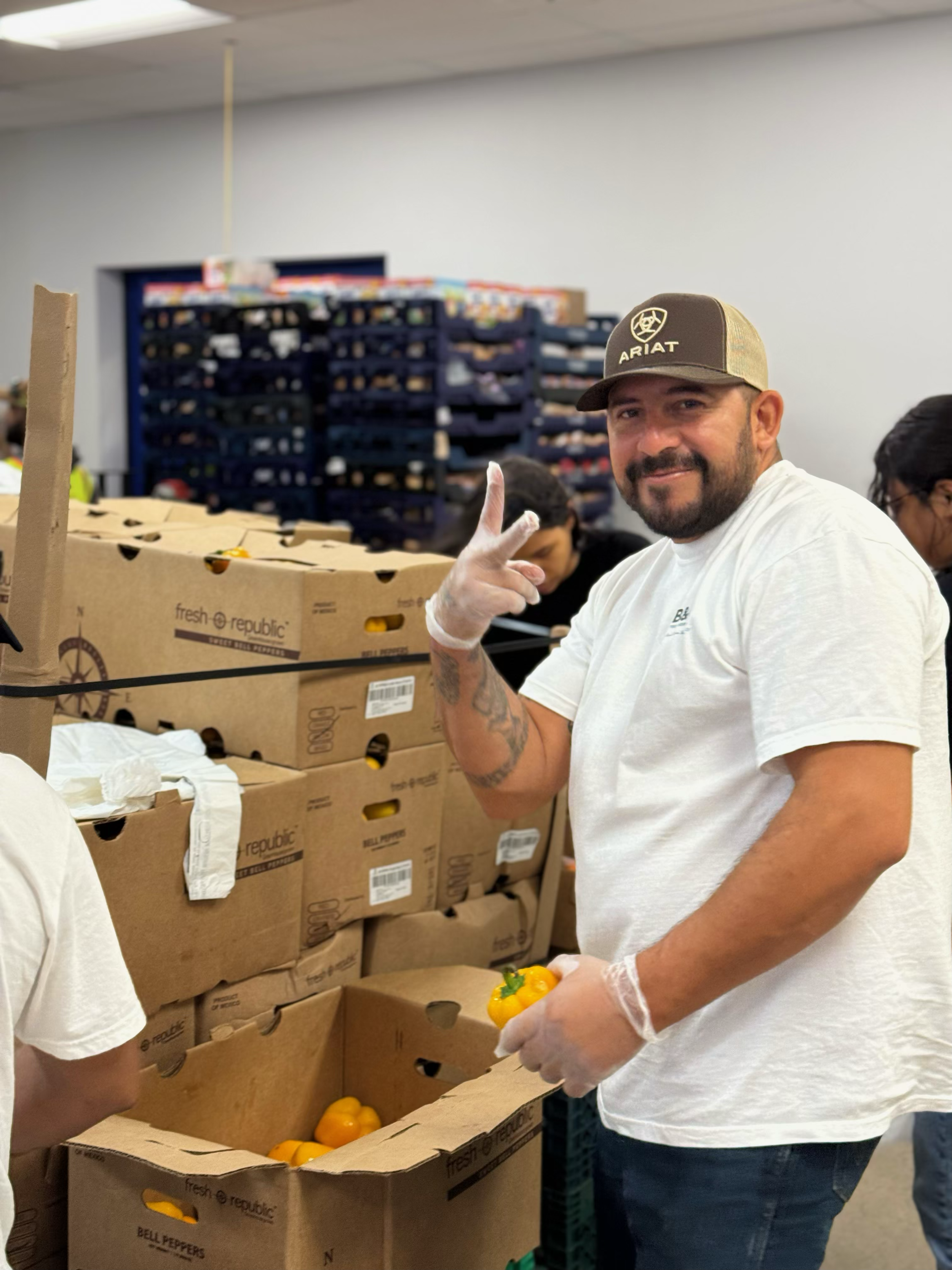 Man wearing a white t-shirt and a baseball cap with the word 'ARIAT' posing and smiling in a warehouse or distribution center, holding a yellow bell pepper, with boxes of bell peppers and other people working in the background.