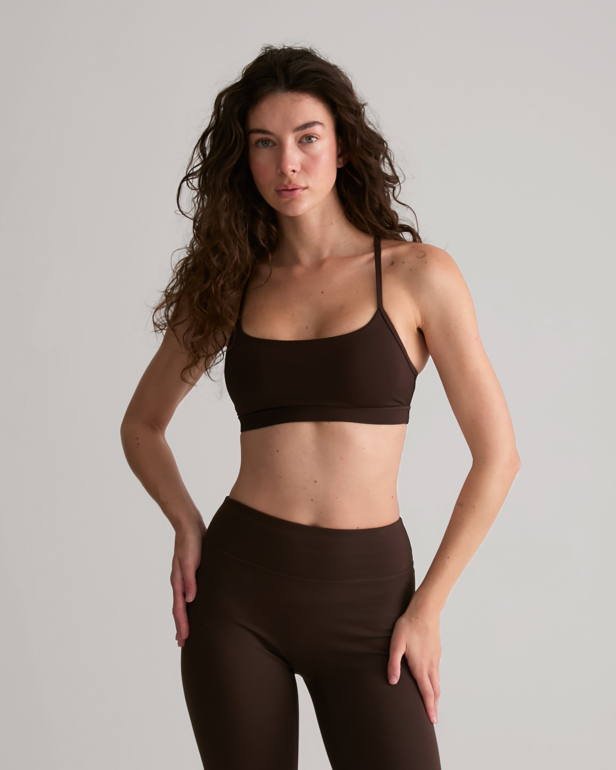 A woman with curly brown hair wearing a black sports bra and black leggings standing against a plain white background.