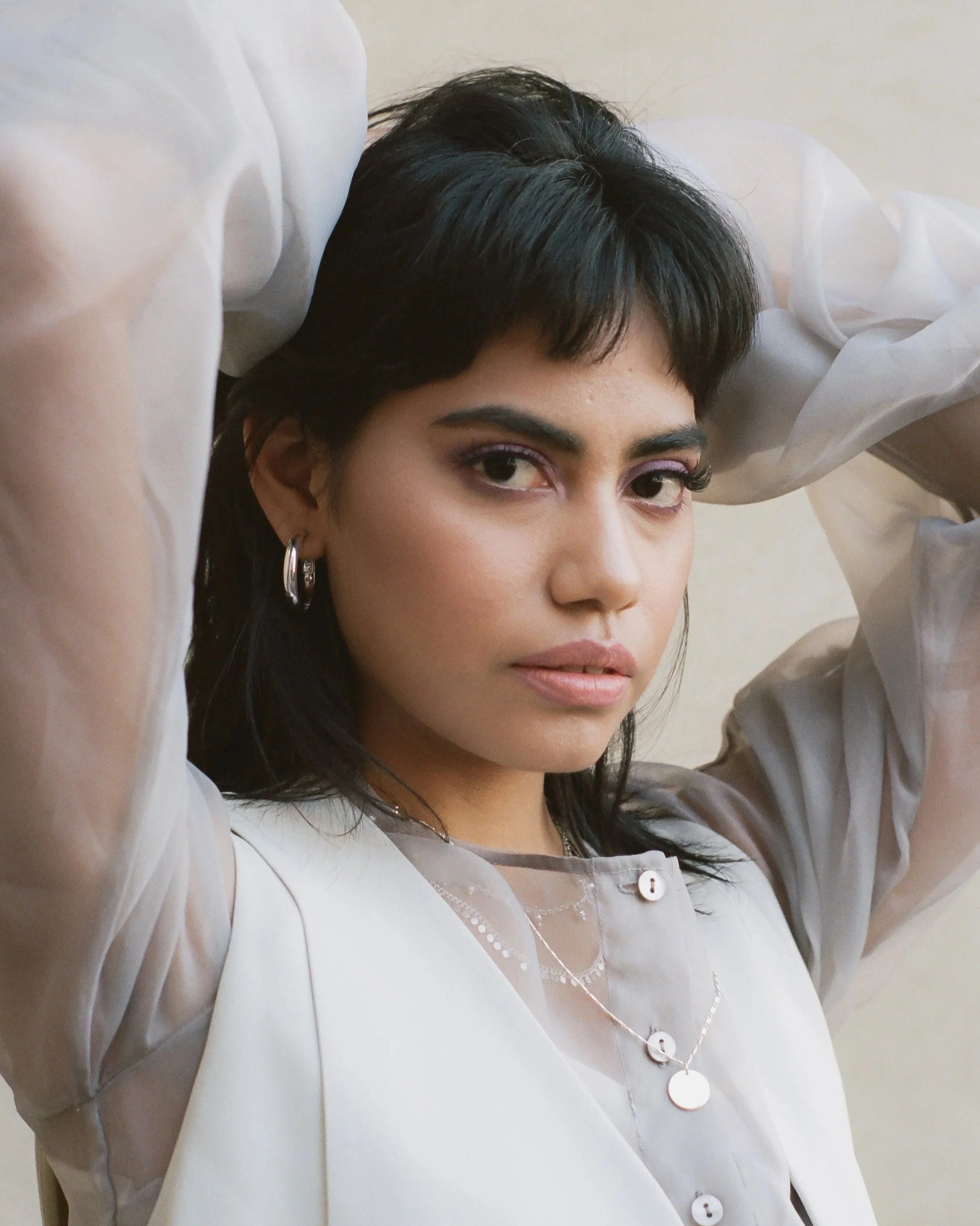 A woman with dark hair is lying down with her arms raised and partly covered by white fabric or clothing. She is wearing earrings and a layered necklace, with makeup accentuating her eyes and lips.