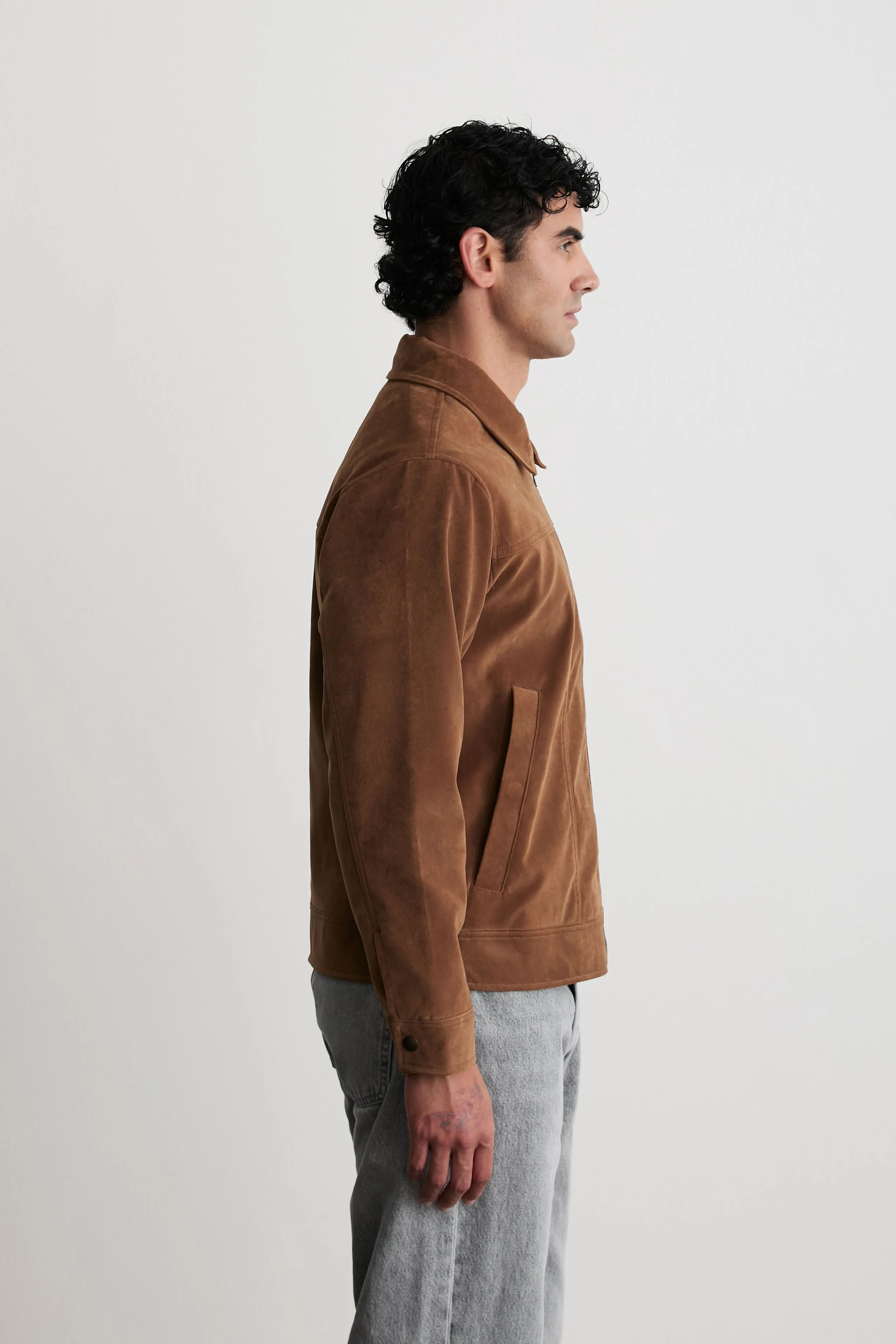 Side view of a man wearing a brown jacket and gray pants, standing against a plain white background.