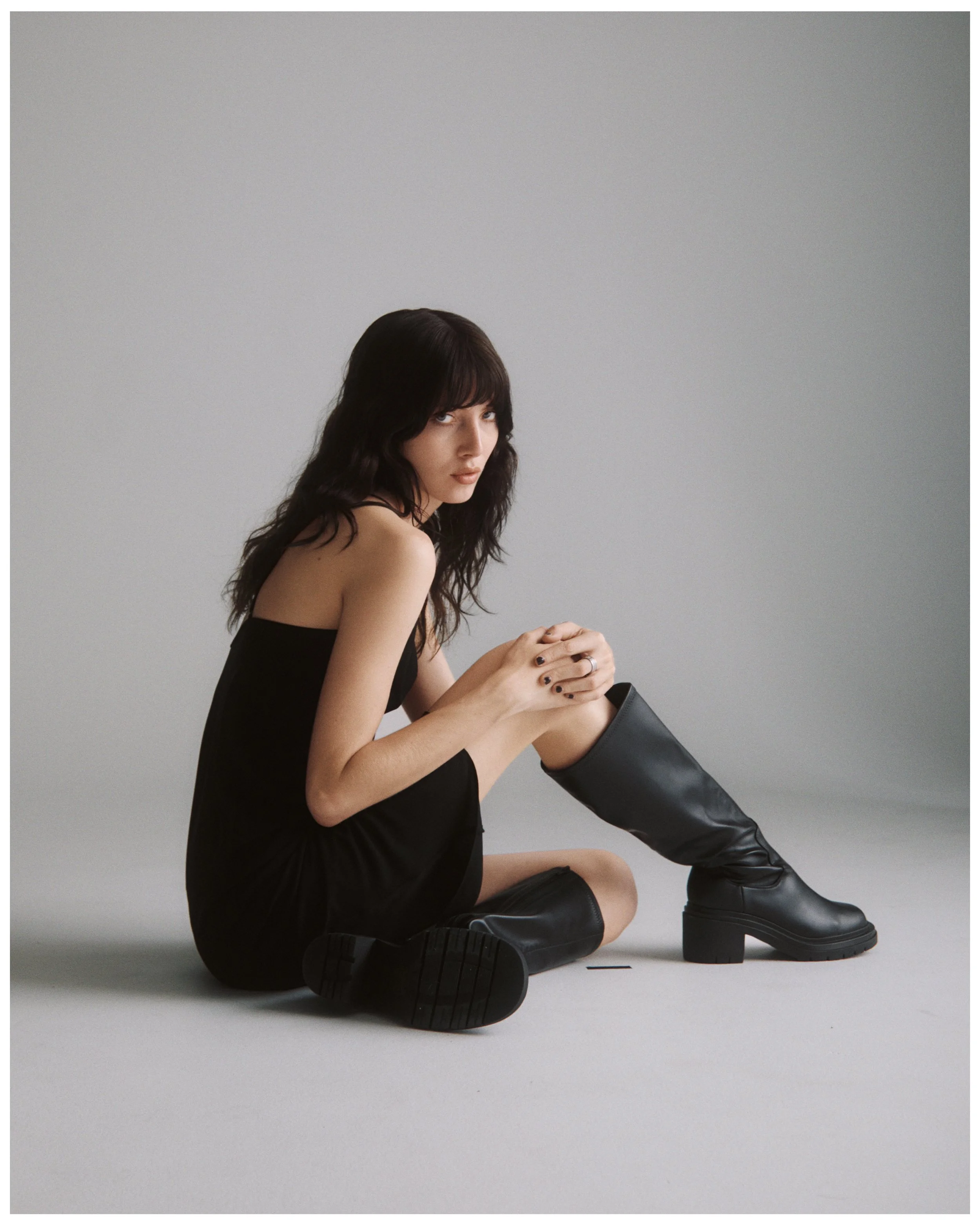 A woman with dark wavy hair and light skin sits on the floor against a plain light gray background. She is dressed in a black strapless dress and wearing tall black boots. She looks towards the camera with an intense expression, her hands clasped in 