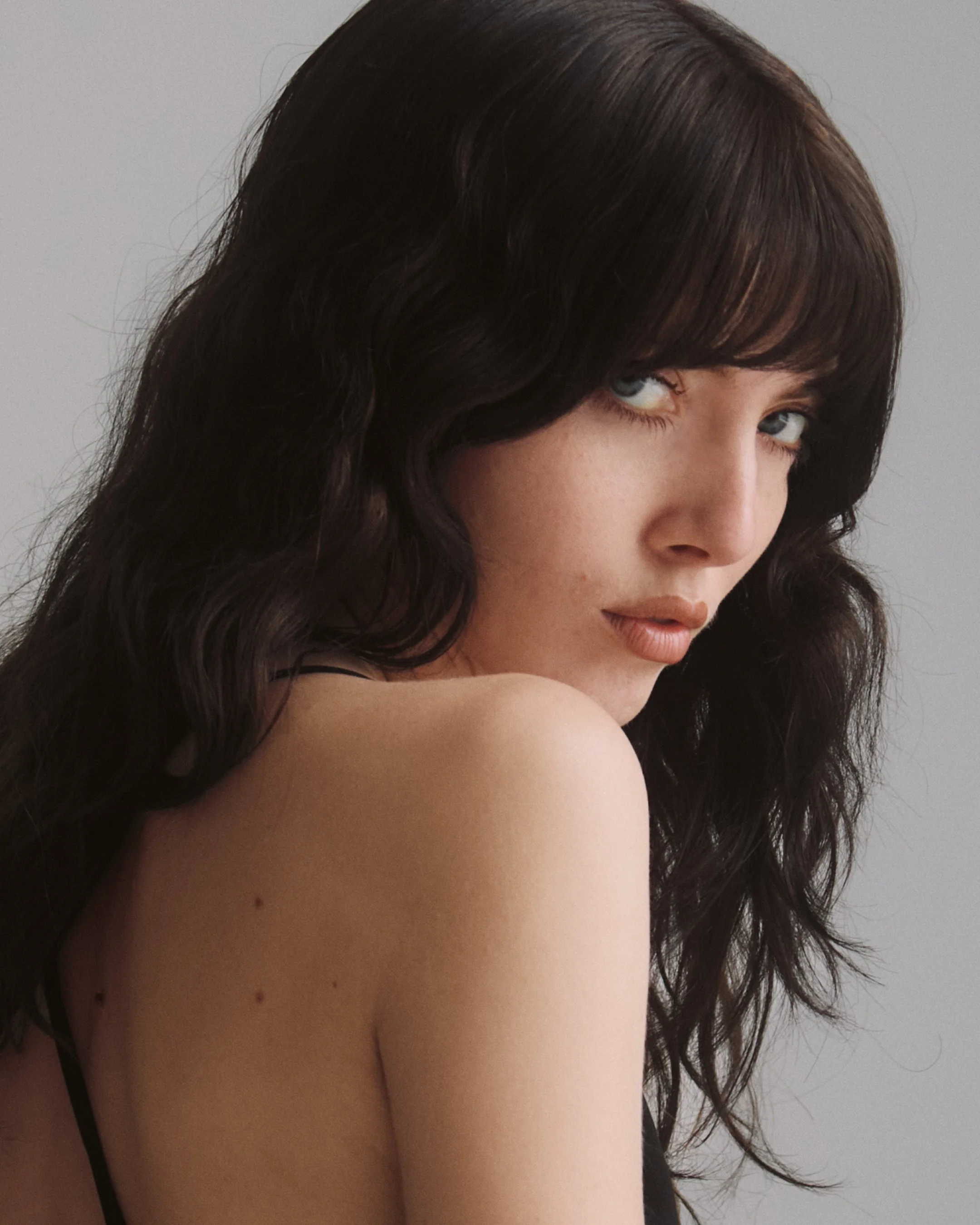 Close-up of a woman with dark, wavy hair and blue eyes, looking over her shoulder, with a neutral gray background.