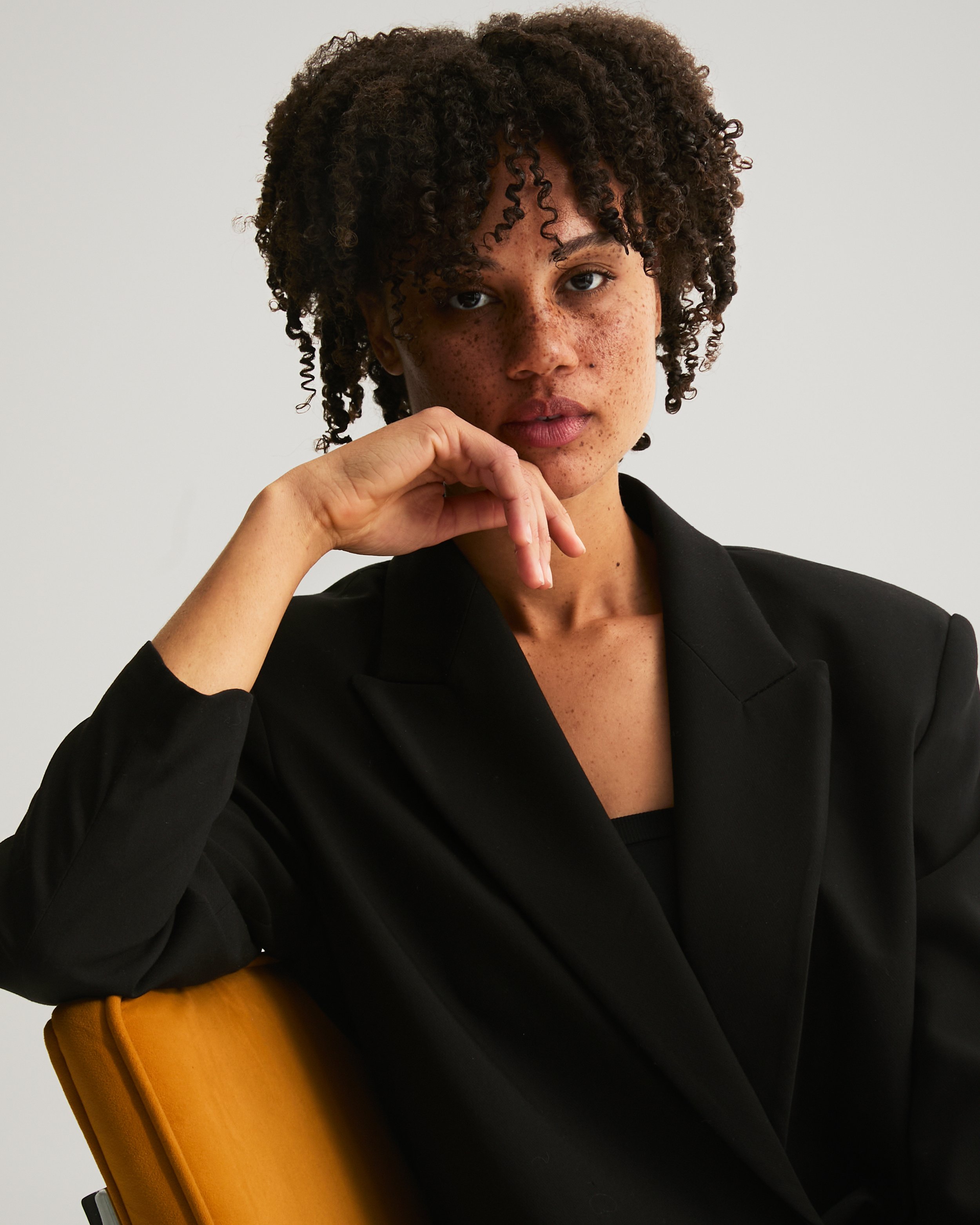 Portrait of a woman with curly hair, wearing a black blazer, sitting on a yellow chair, with her hand near her face, against a plain white background.
