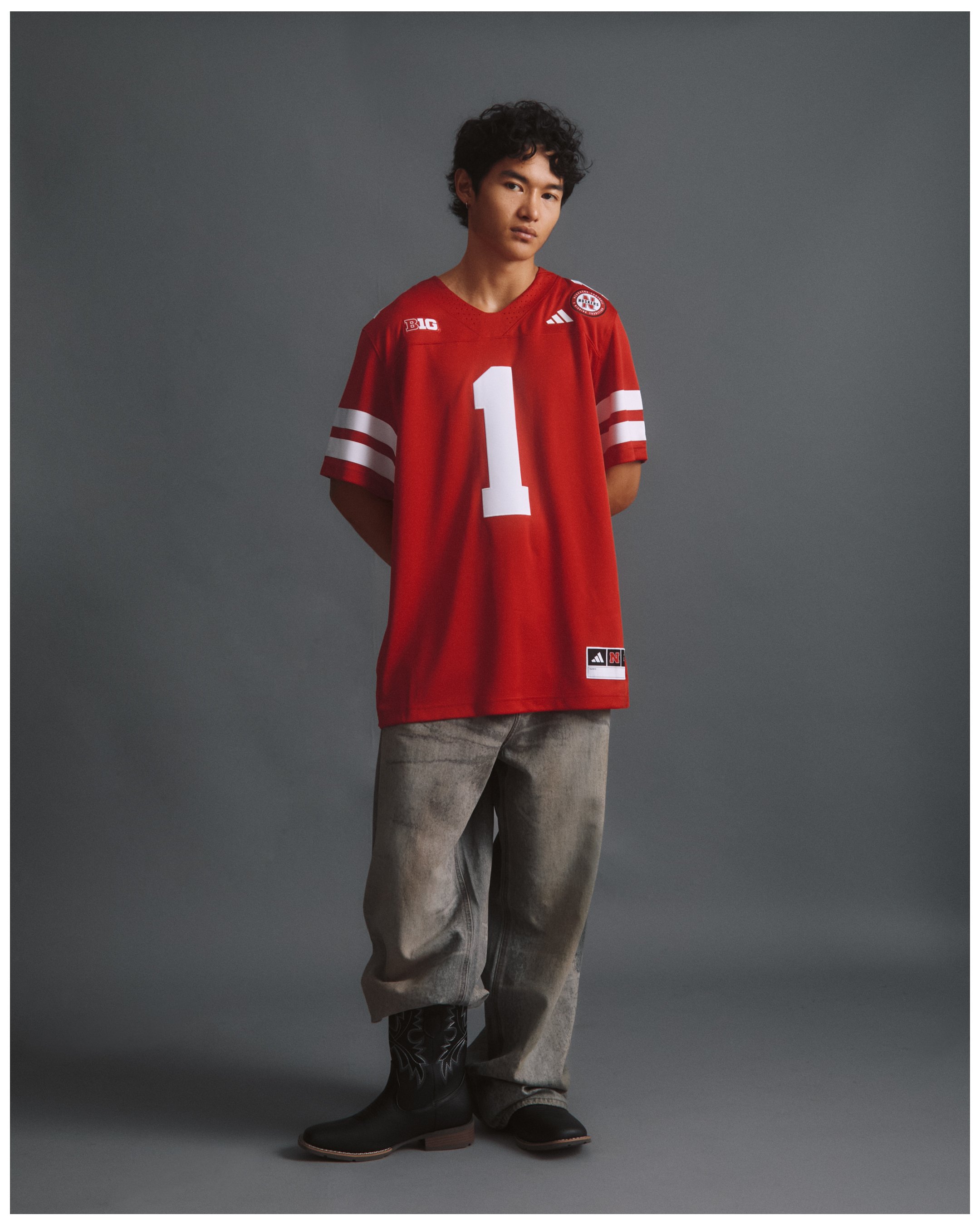 A person wearing a red sports jersey with the number 1, paired with gray pants, patterned black boots, and standing against a gray backdrop.