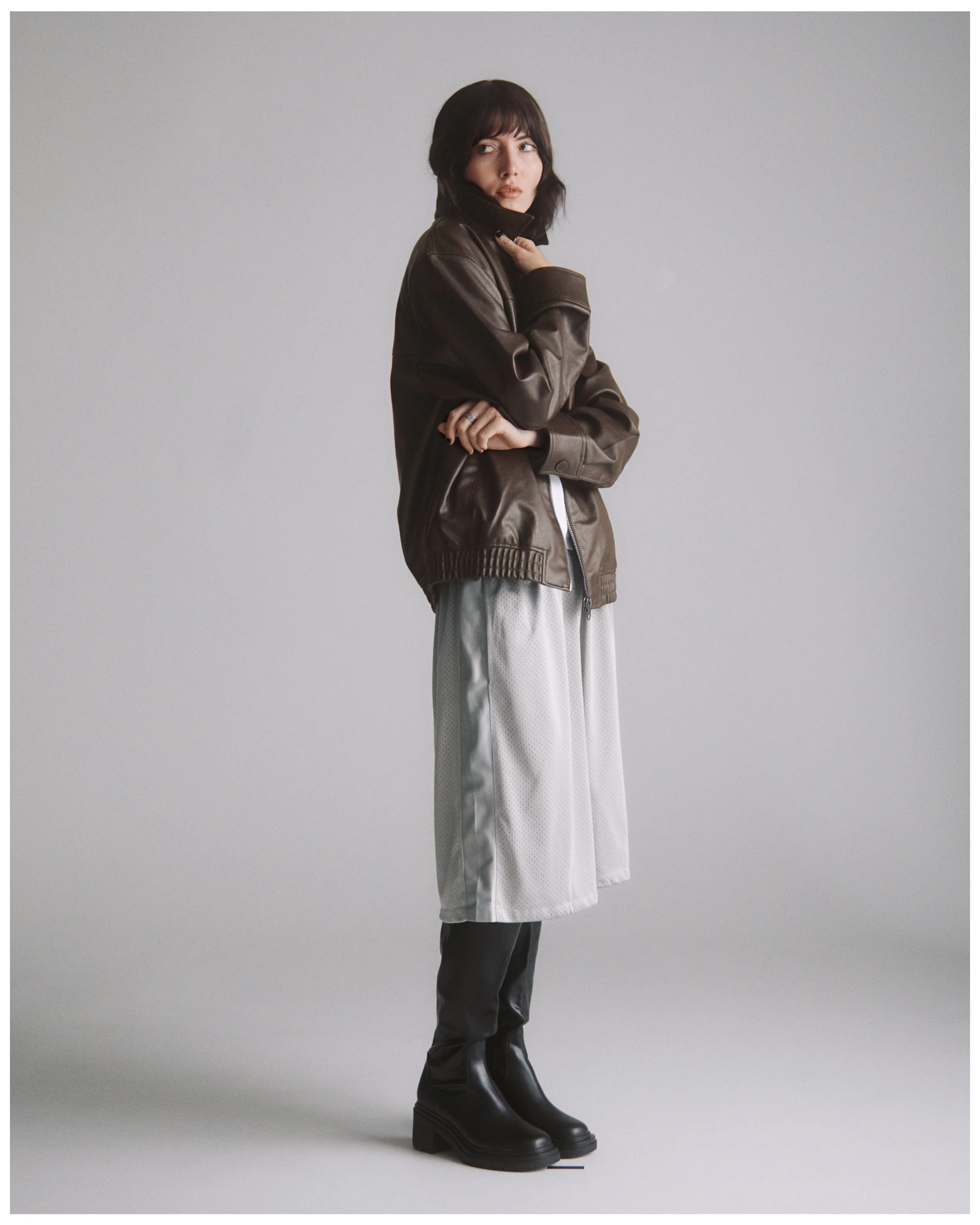 Fashion model wearing a brown leather jacket, light-colored skirt, black tights, and chunky black boots, standing against a plain gray background.