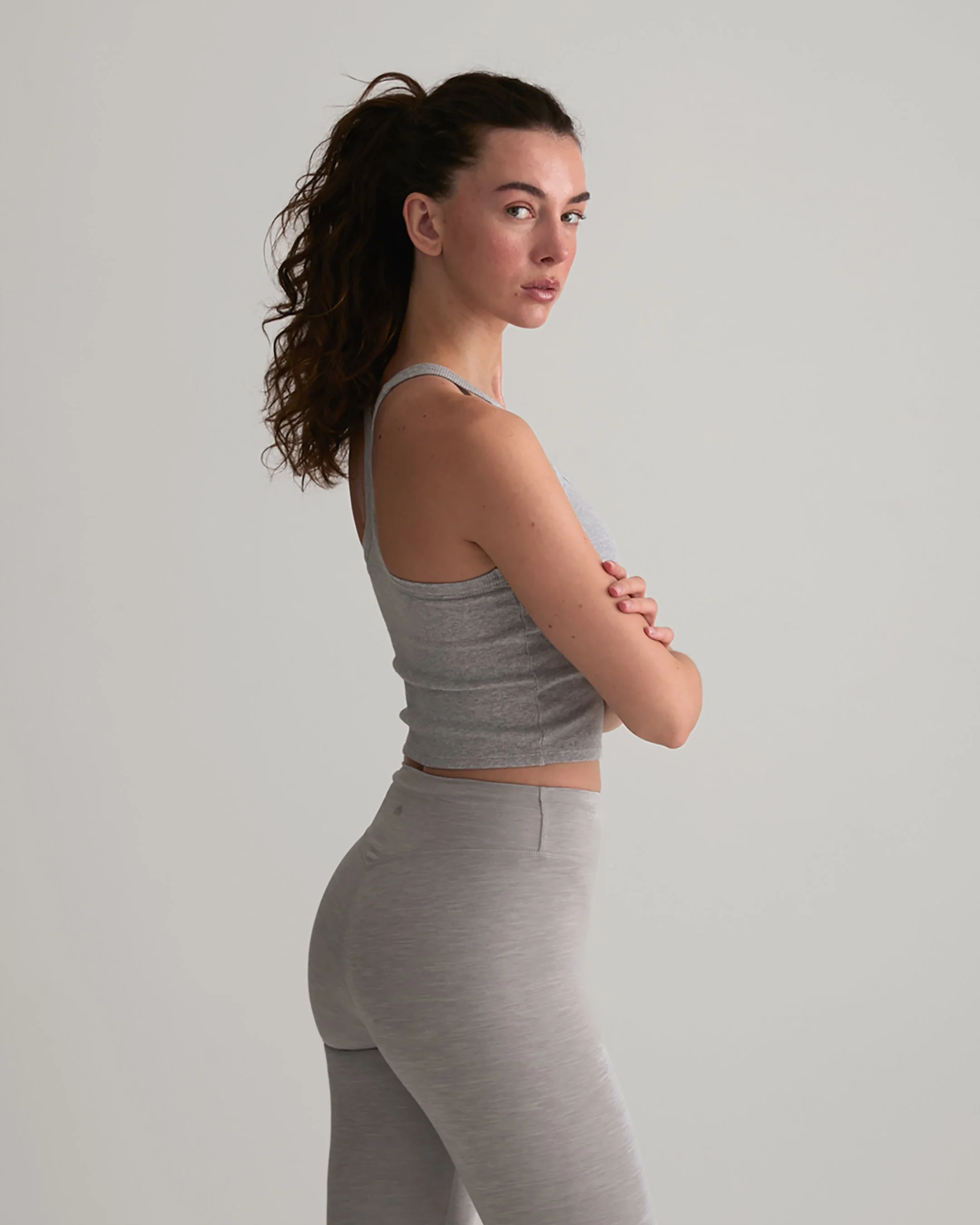 A woman with curly brown hair standing against a plain white background. She is wearing a gray tank top and matching gray leggings, with her arms crossed and looking at the camera.