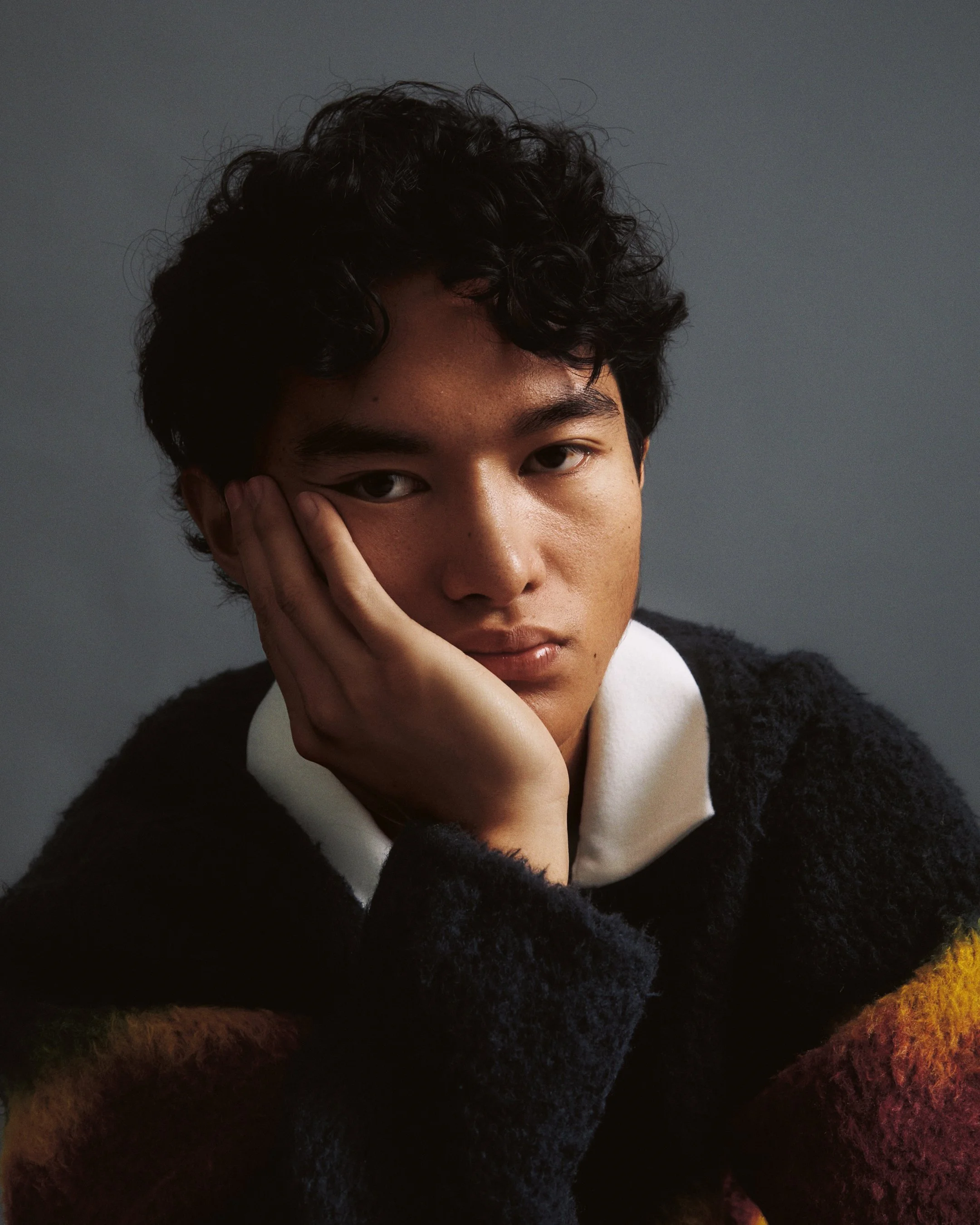 A young person with black curly hair and tan skin, resting their face on their hand, looking at the camera in a close-up portrait against a neutral background.