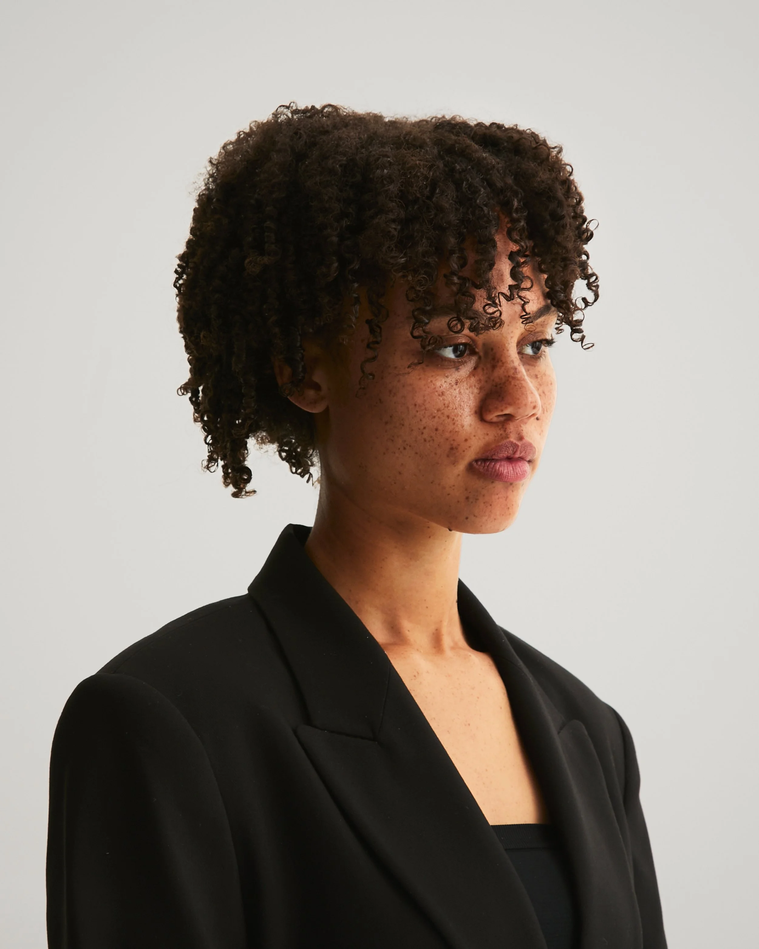A woman with curly brown hair, freckles, and wearing a black blazer, looking to her right against a plain light gray background.