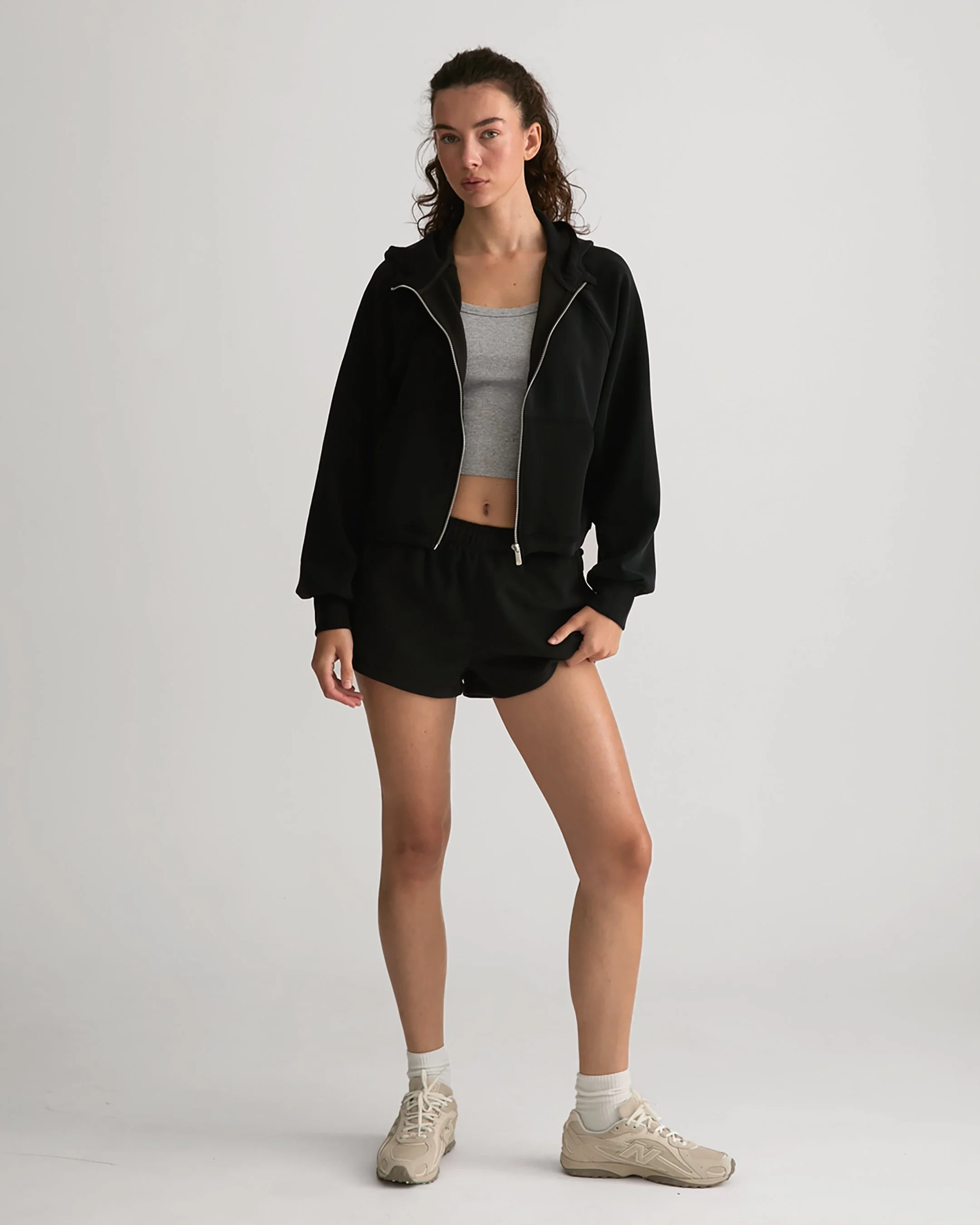 A young woman wearing beige sneakers, white socks, black shorts, a gray crop top, and a black zip-up athletic jacket standing against a plain white background.