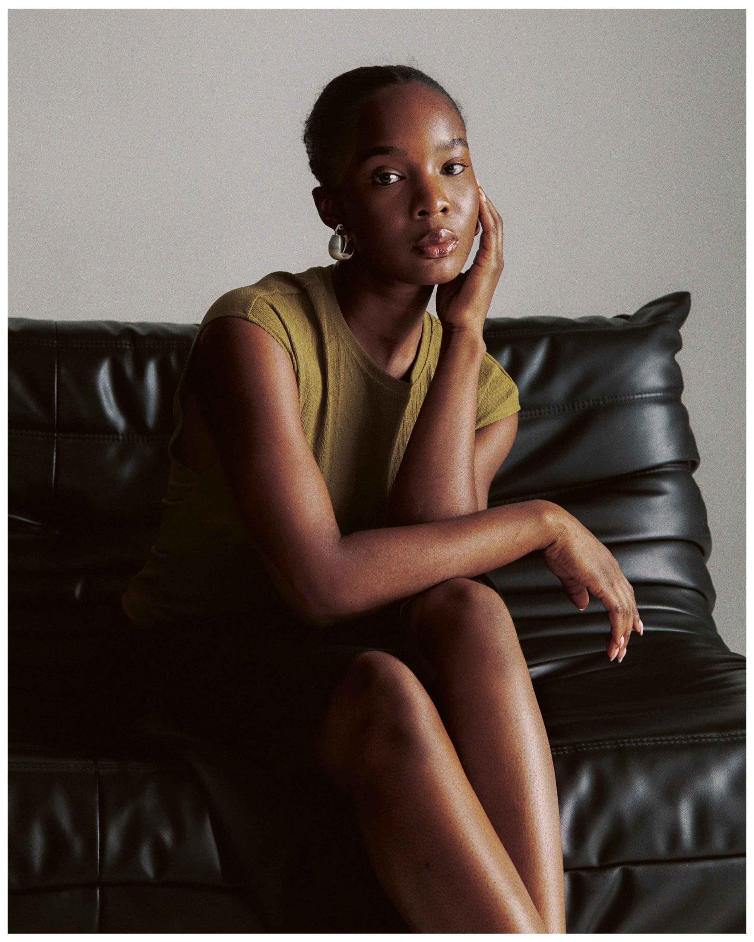 A portrait of a young Black woman with short hair, wearing a light green T-shirt and silver earrings, sitting on a black leather sofa against a plain light gray background.