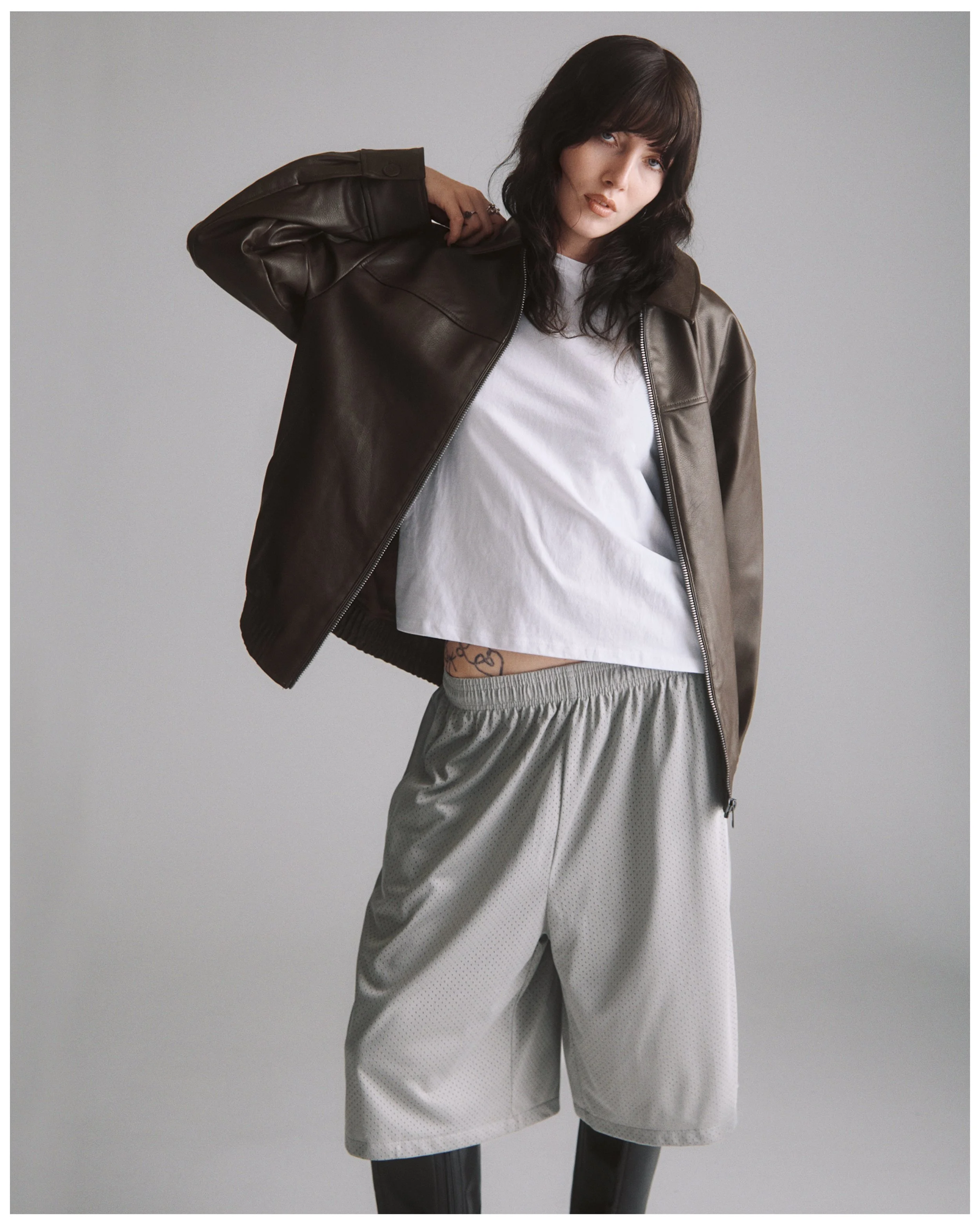 Woman with dark hair and bangs wearing a white T-shirt, grey sweatpants, black leggings, and a dark brown leather jacket, standing against a light grey background.