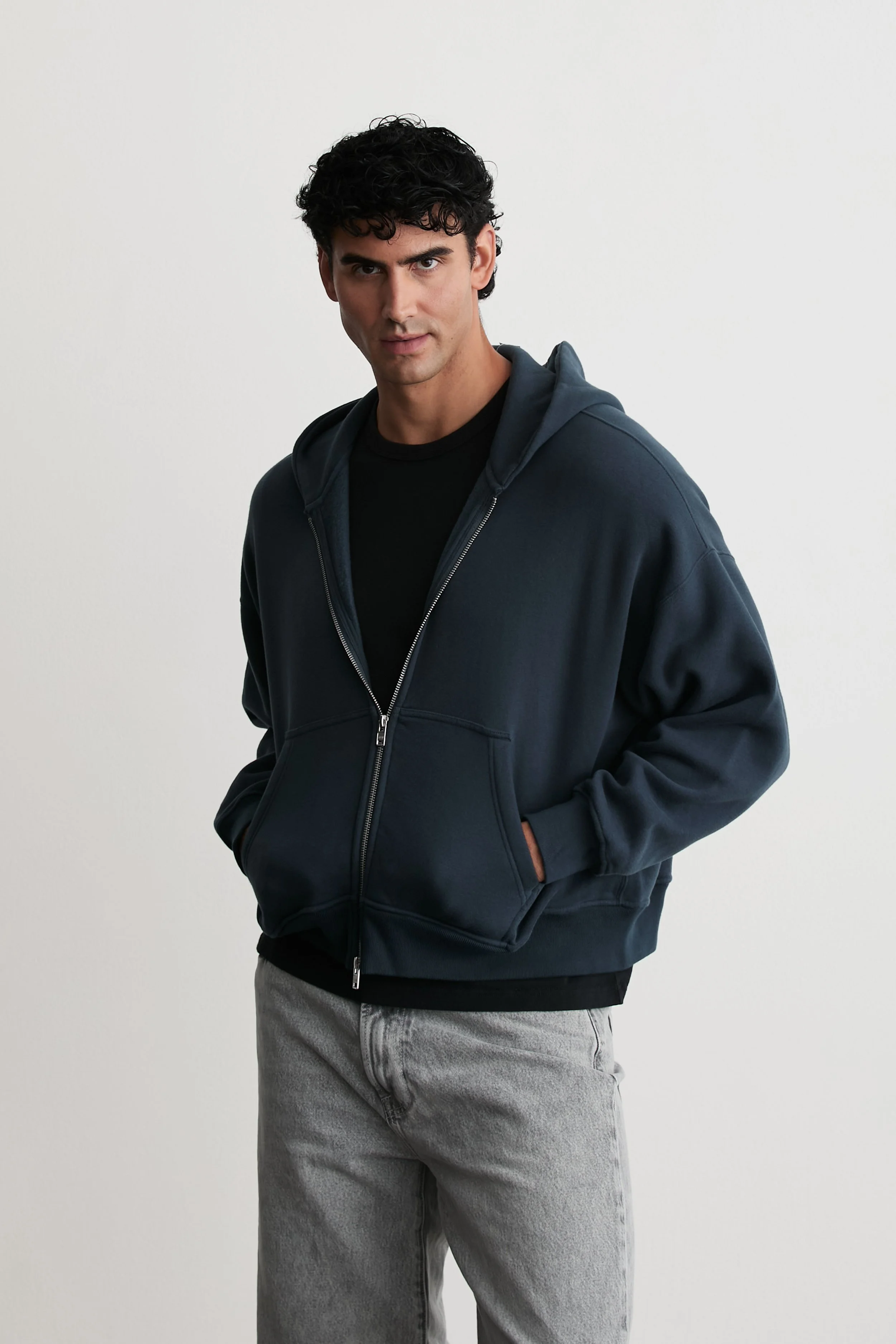 A young man with curly black hair wearing a dark hoodie over a black t-shirt and light gray jeans, standing against a plain white background.