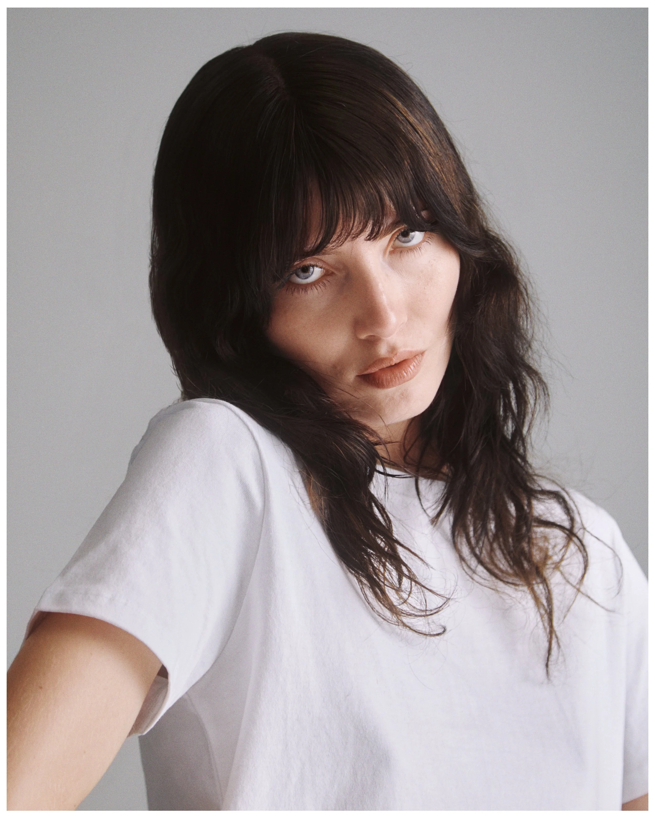 A woman with dark, long wavy hair and fair skin wearing a white shirt, standing against a plain light gray background.