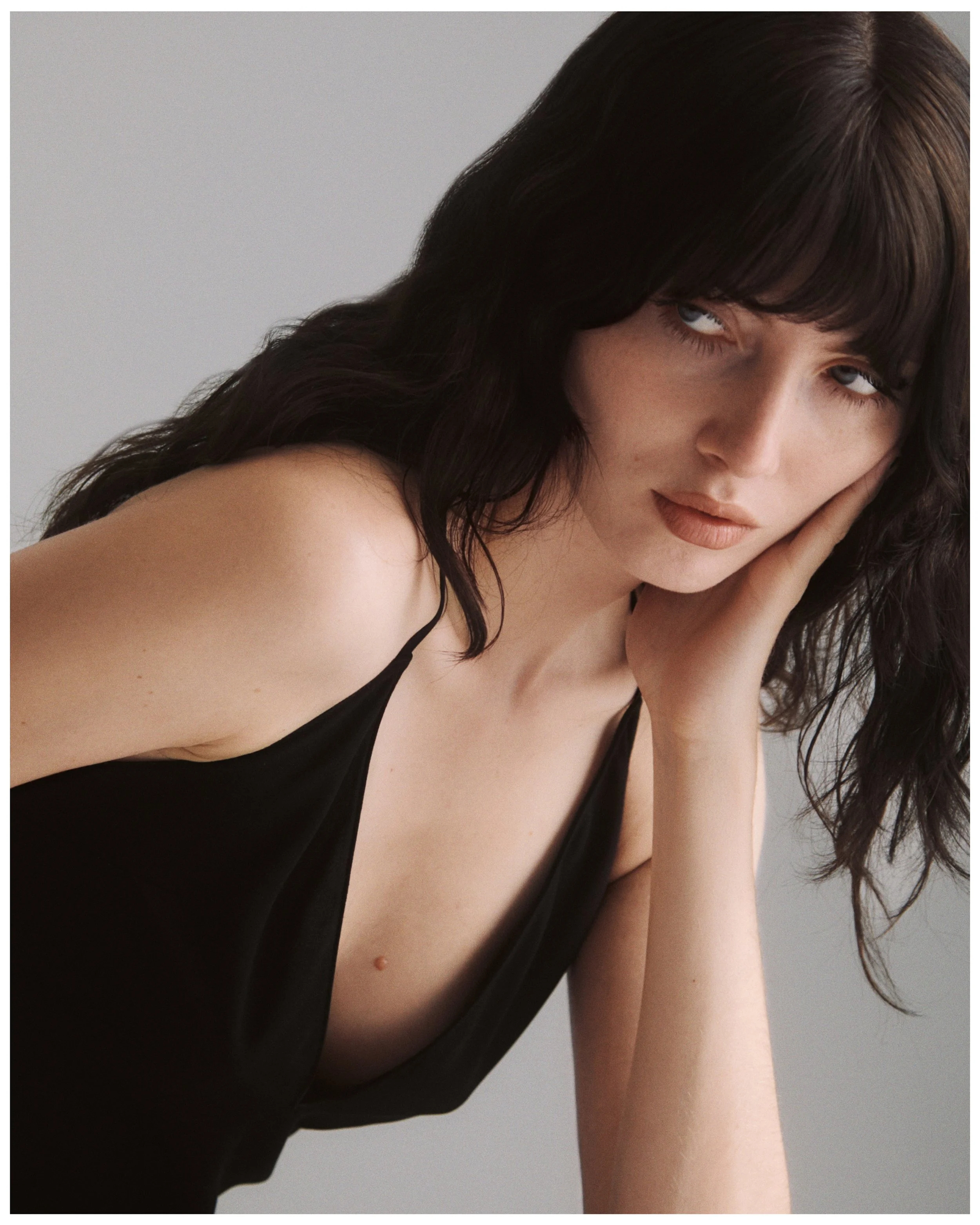 A woman with dark wavy hair and blue eyes wearing a black spaghetti strap top, resting her face on her hand, with neutral background.