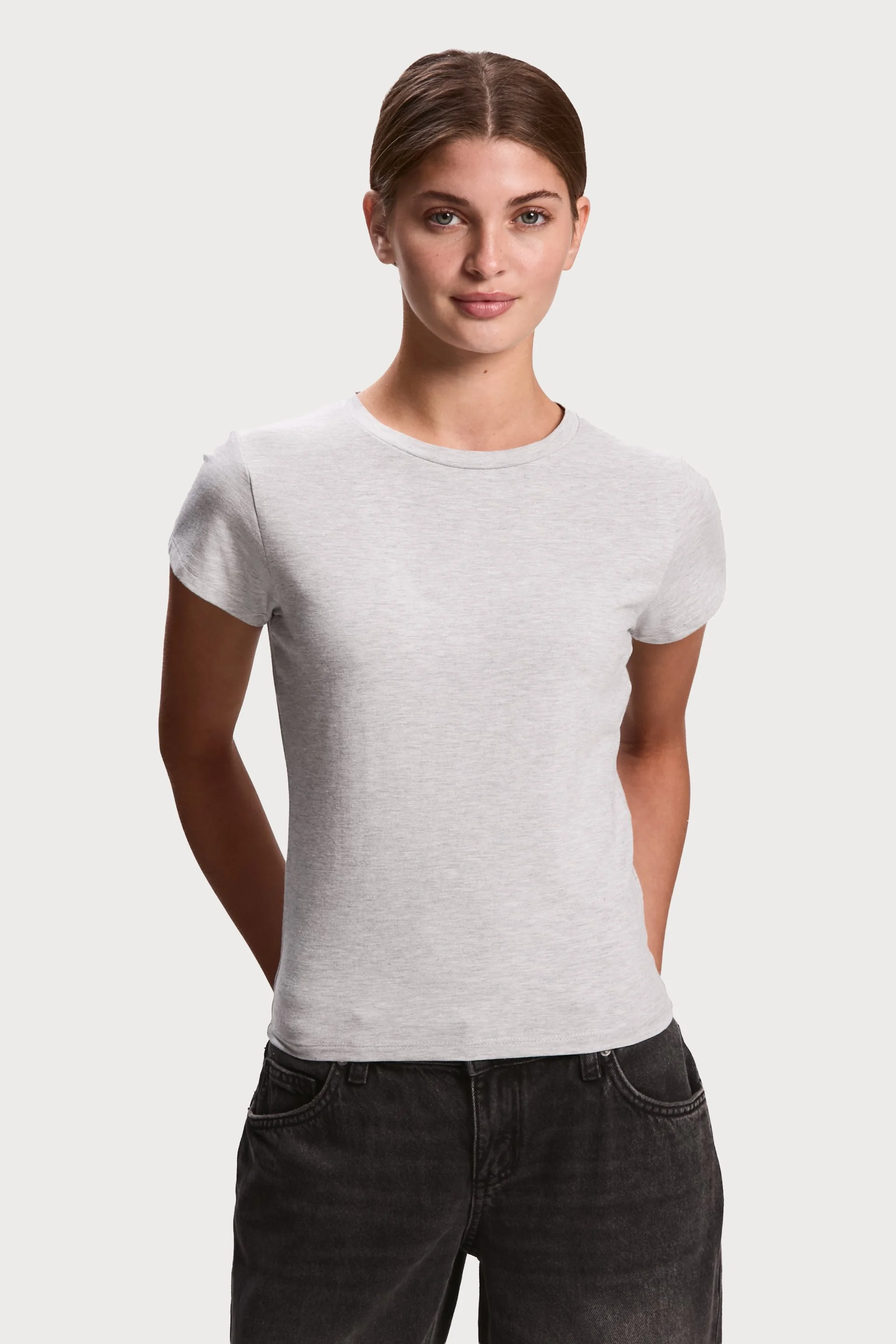 Young woman with brown hair wearing a light grey t-shirt and black jeans, standing against a light grey background.