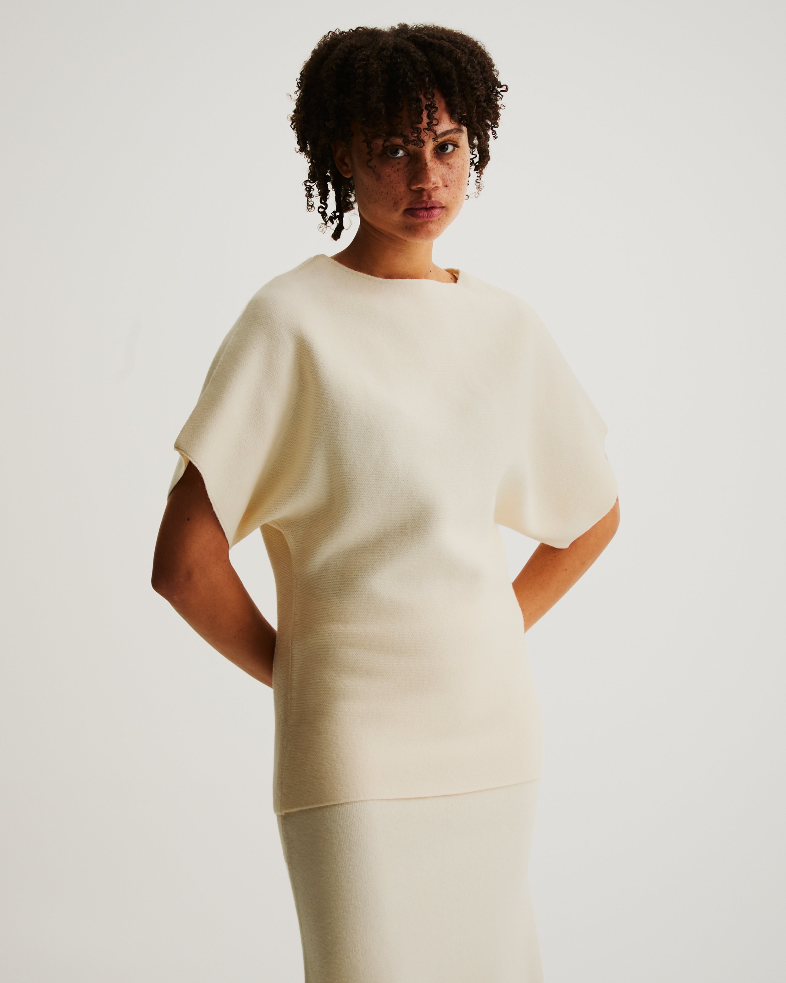 A woman with curly hair wearing a cream-colored, short-sleeved knit top and matching skirt standing against a plain white background.