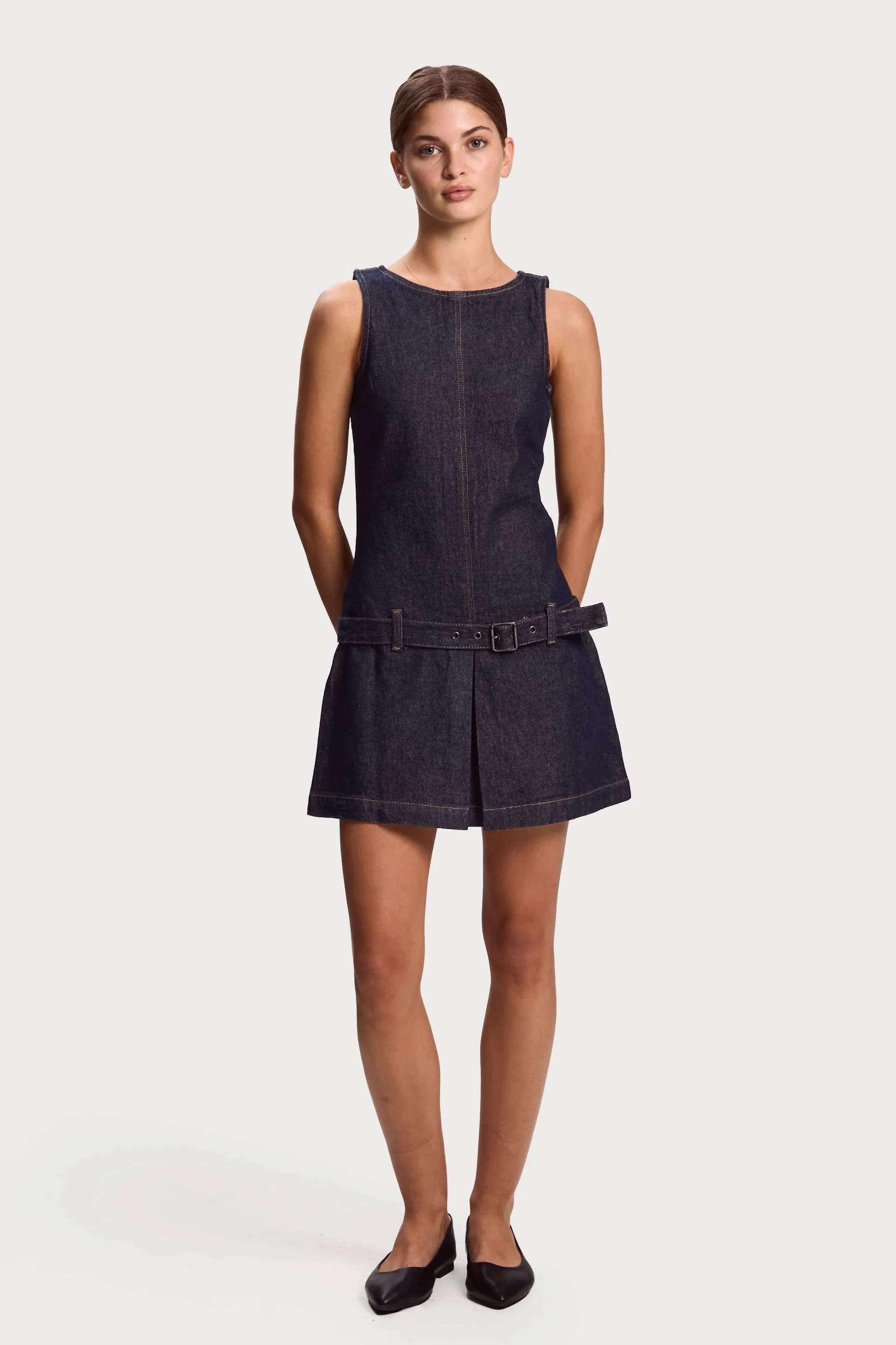 A young woman wearing a sleeveless dark denim dress with a belt and black flat shoes, standing against a plain light gray background.