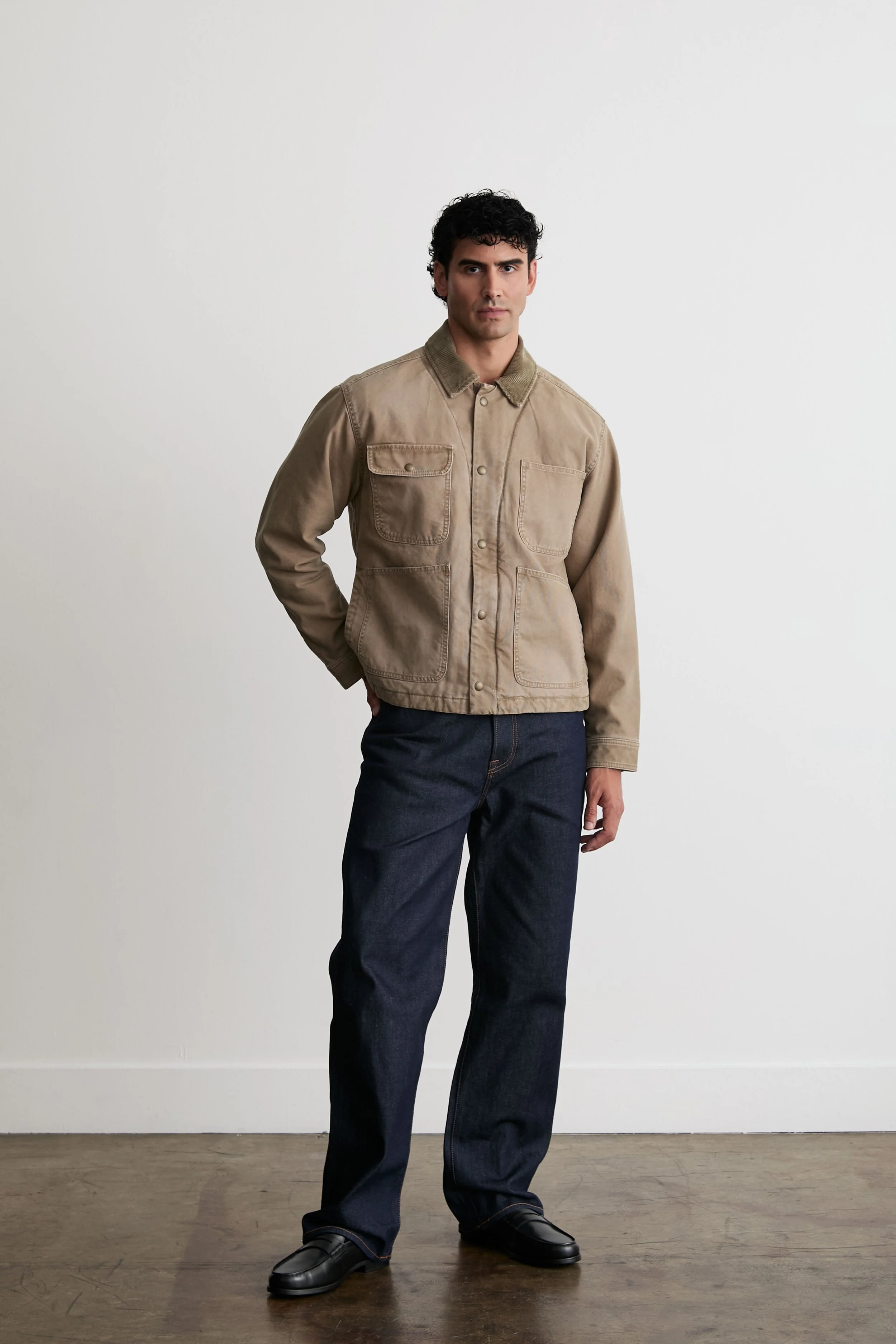 A man standing indoors against a plain white wall, wearing a beige oversized jacket with large front pockets, dark blue jeans, and black shoes, looking slightly to the side.