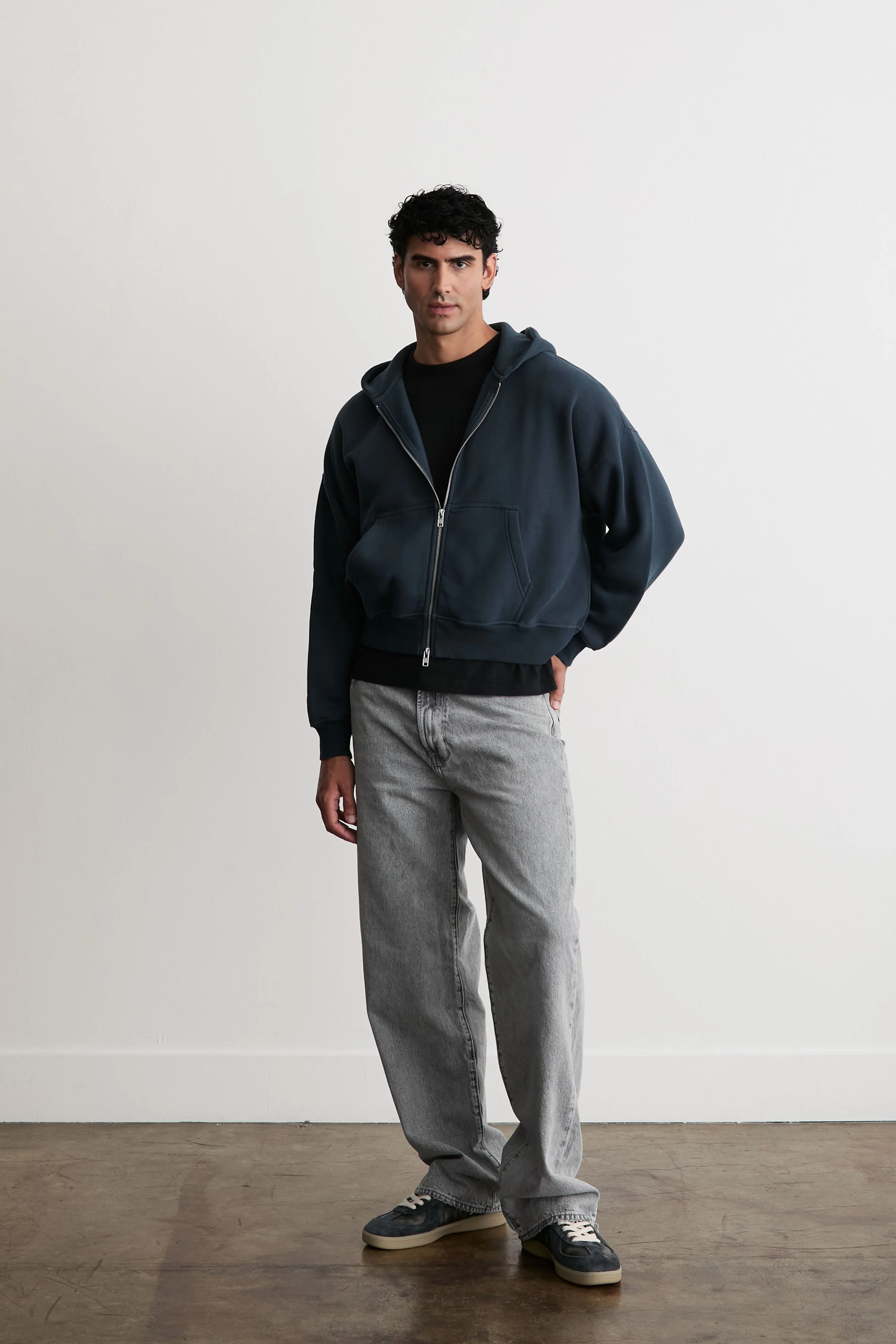 Young man with dark curly hair wearing a navy hoodie, black t-shirt, gray jeans, and sneakers, standing against a plain white wall.