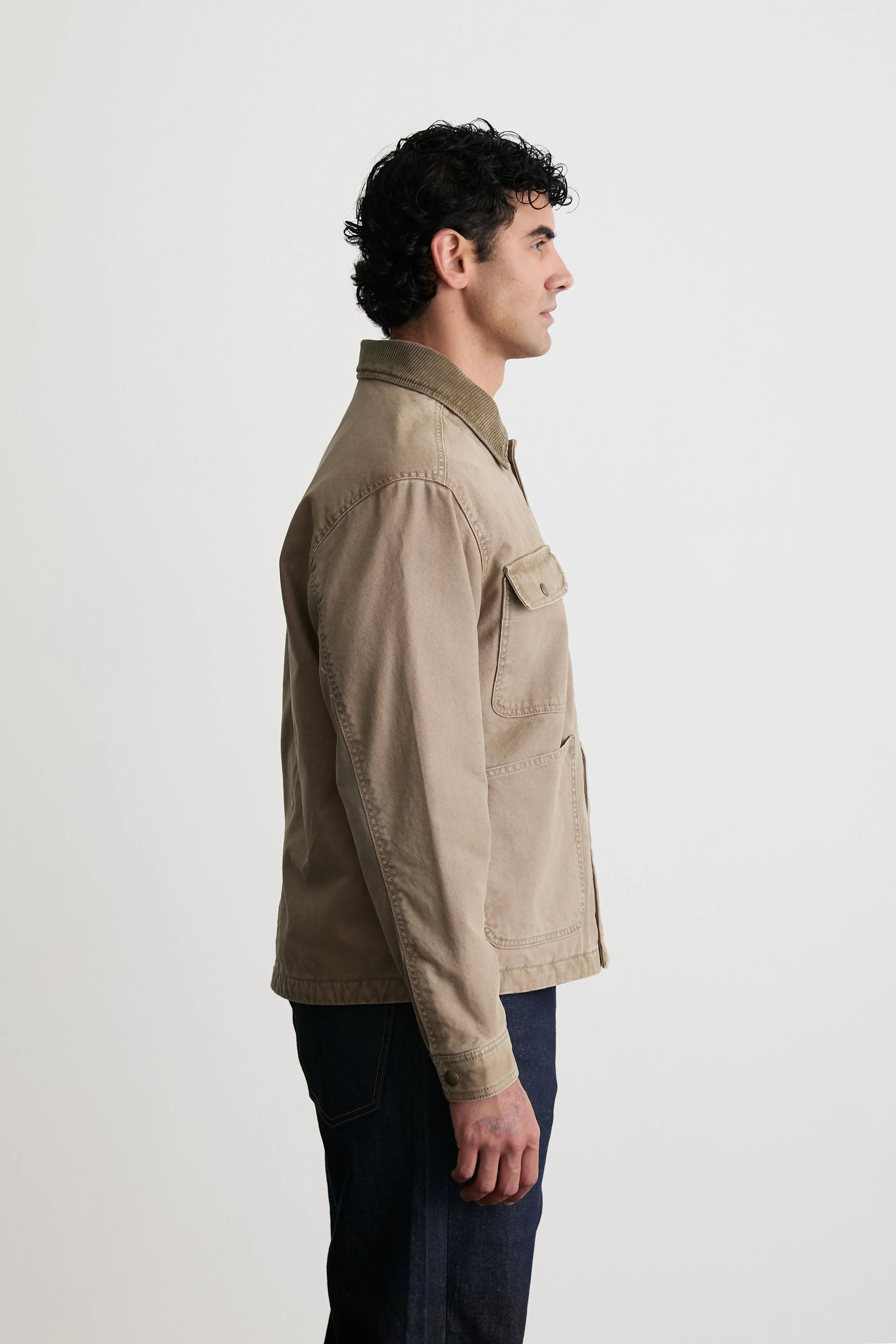 Side view of a man with dark curly hair wearing a beige jacket with a tan collar, dark jeans, standing against a plain white background.