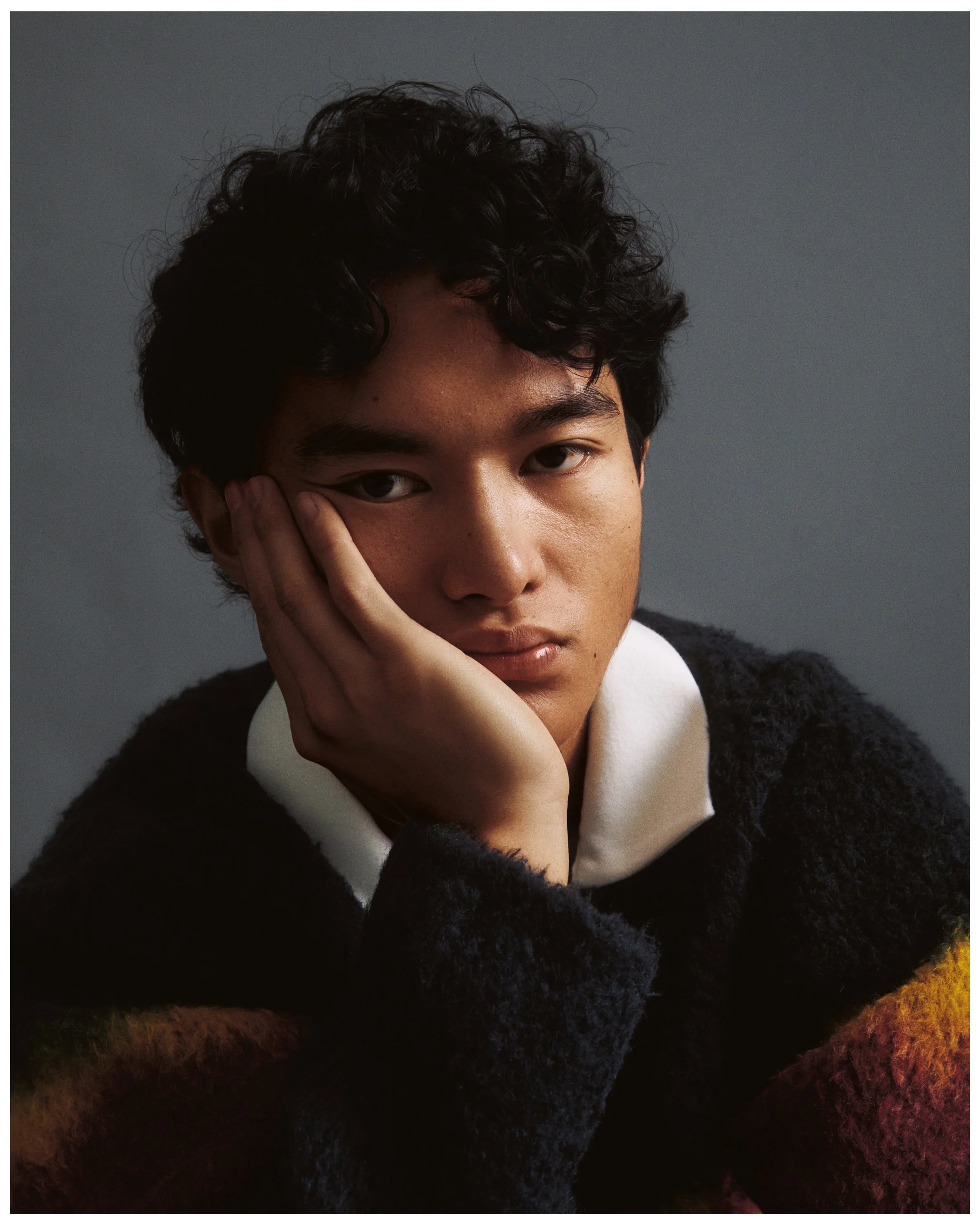 A close-up portrait of a person with short curly dark hair resting their face on their hand against a gray background. They are wearing a black textured sweater and have a calm, neutral expression.