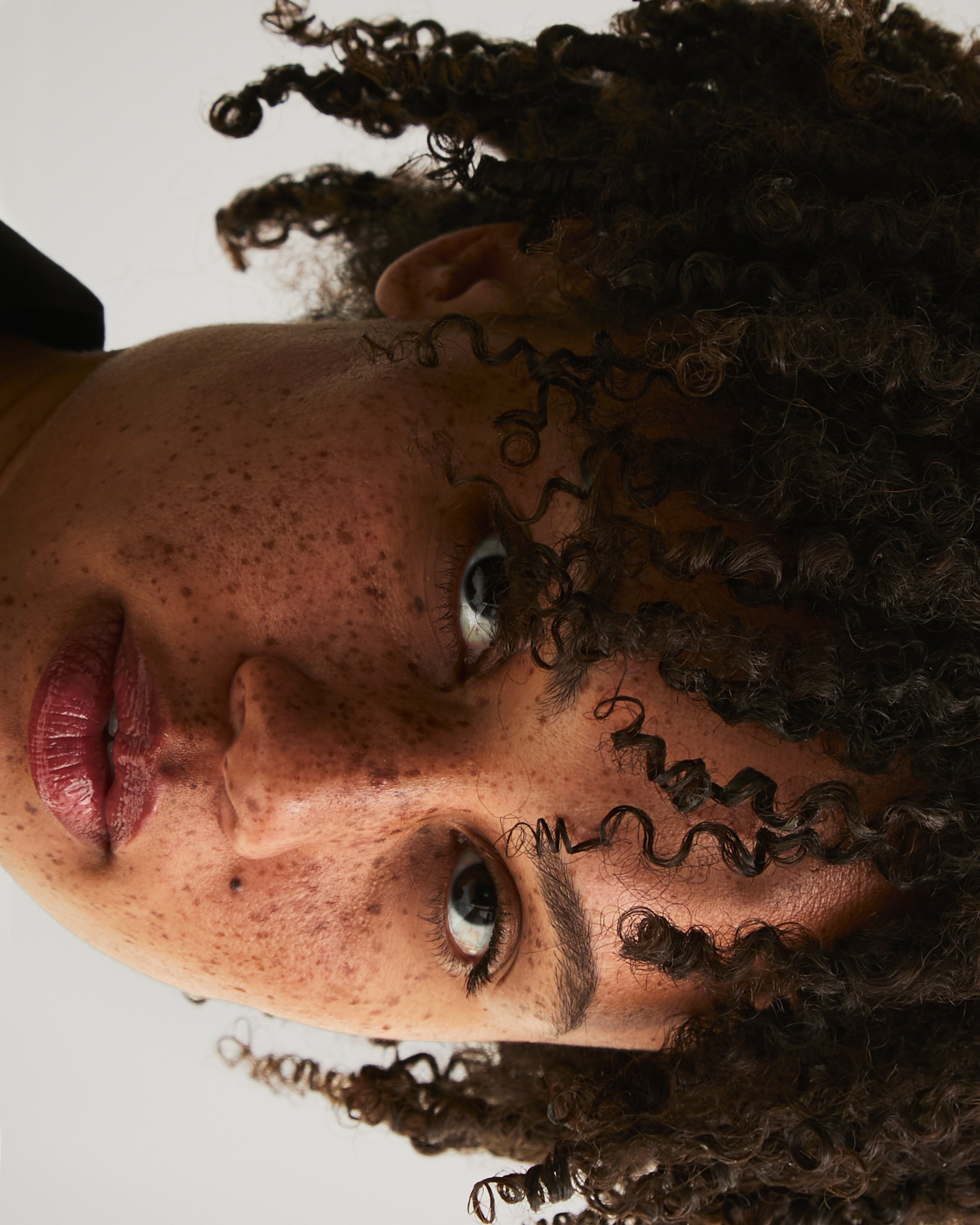 A close-up portrait of a woman with curly hair, freckles, and light makeup, looking directly at the camera against a white background.