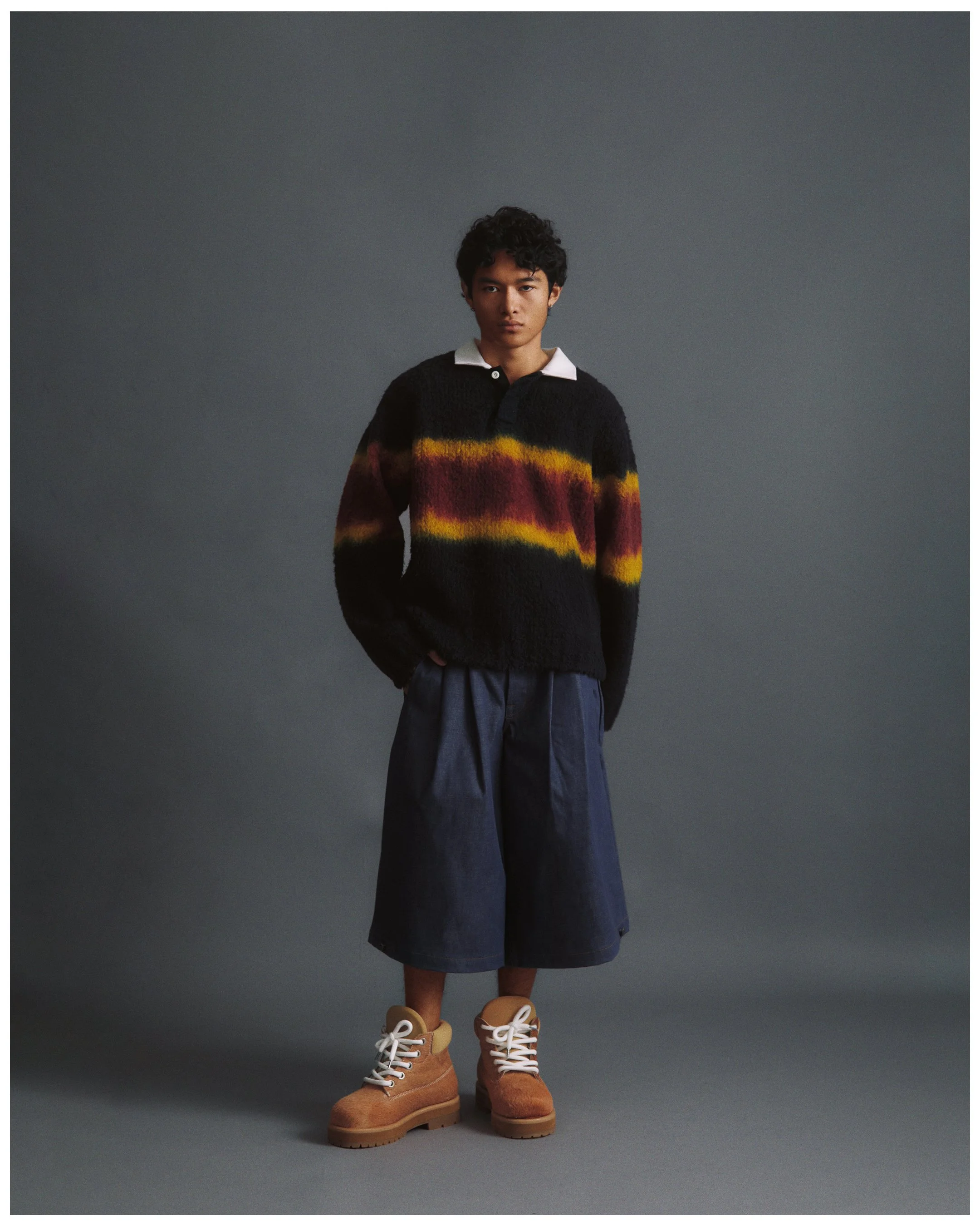 A person with a neutral expression wearing a multi-colored sweater, loose dark shorts, tan boots, and beige ankle socks, standing against a plain grey background.