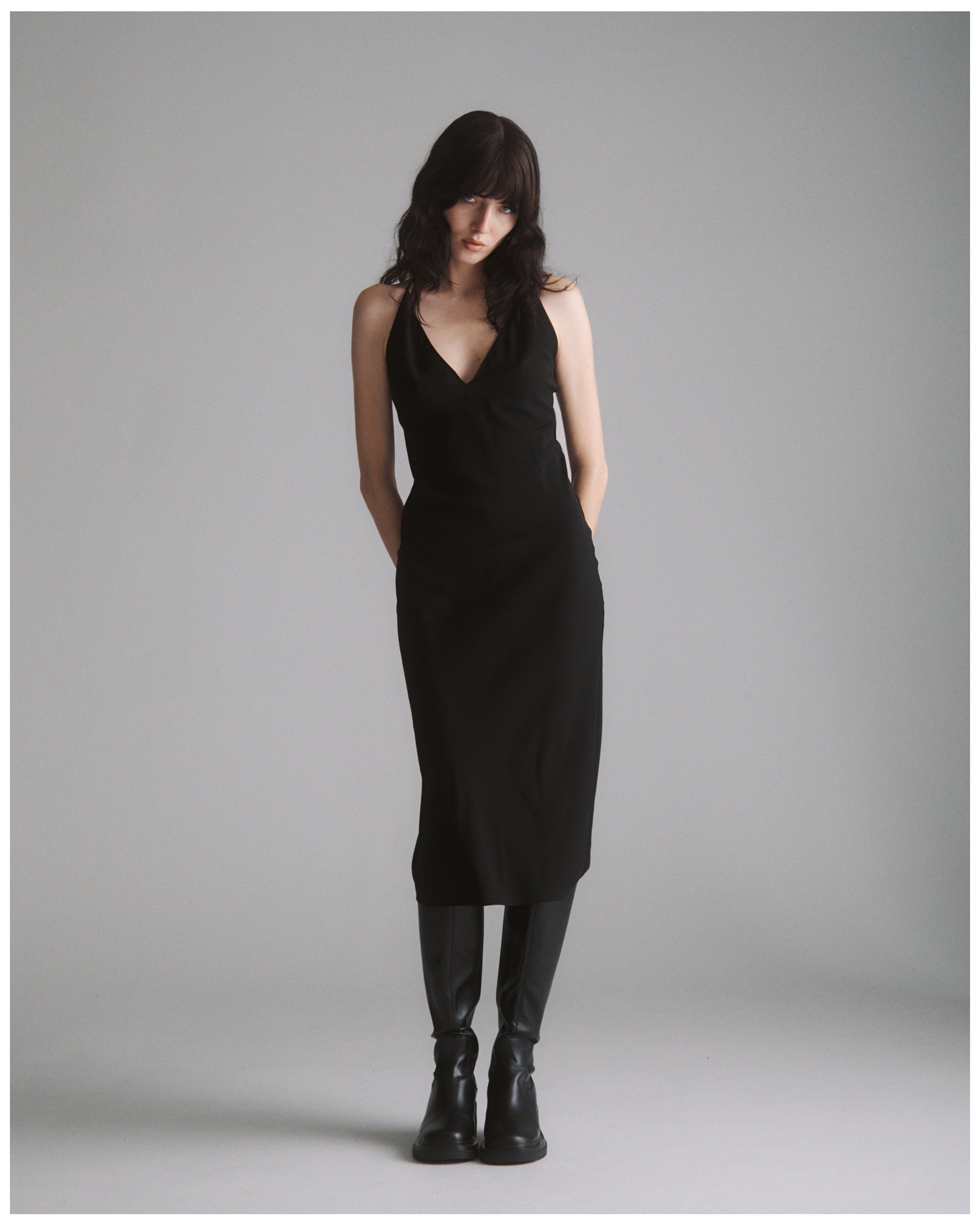A woman with dark hair, wearing a sleeveless black dress, stands against a plain, light gray background, with her hands behind her back and looking at the camera.