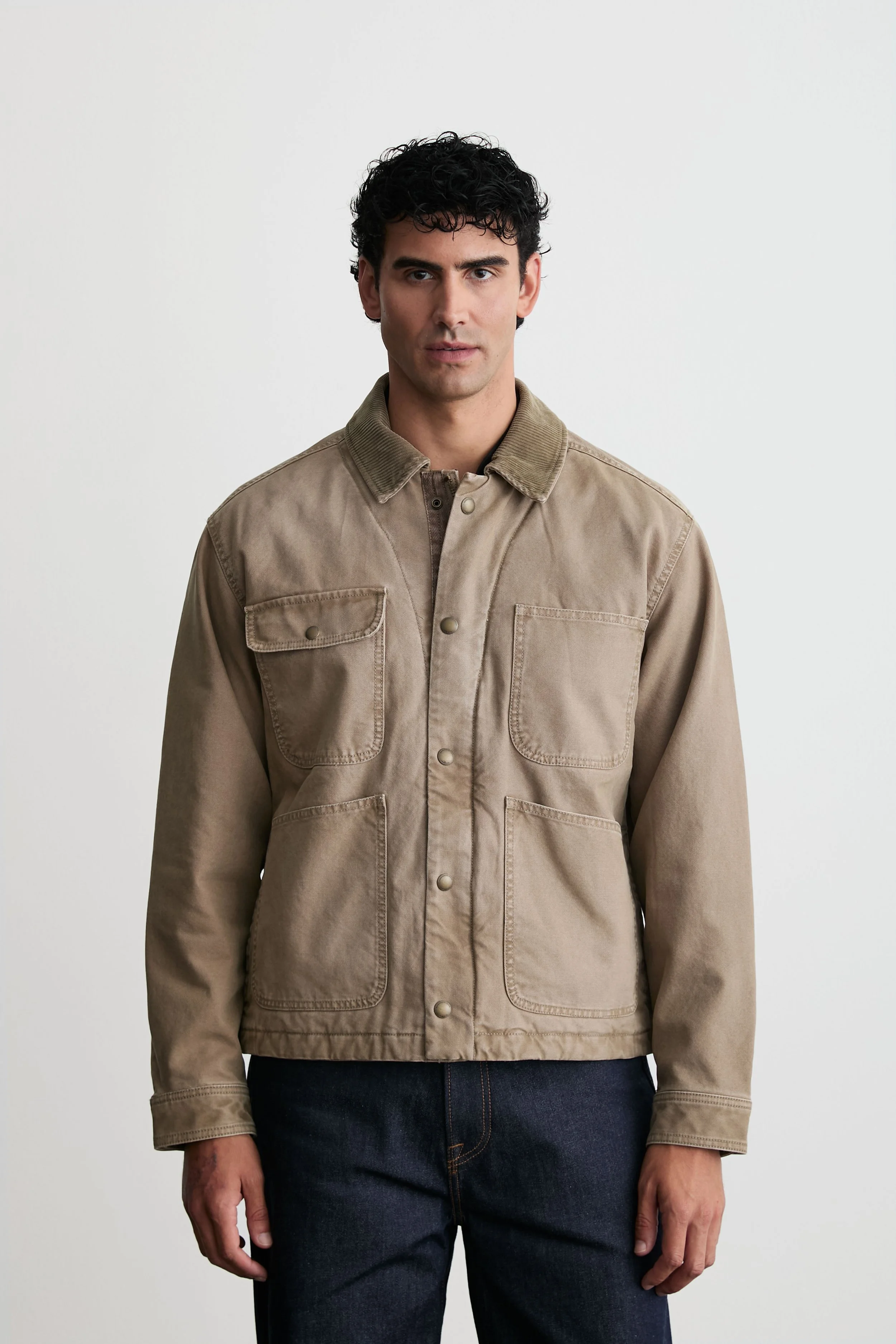 A man with short, curly black hair wearing a tan button-up jacket and dark jeans, standing against a plain white background.