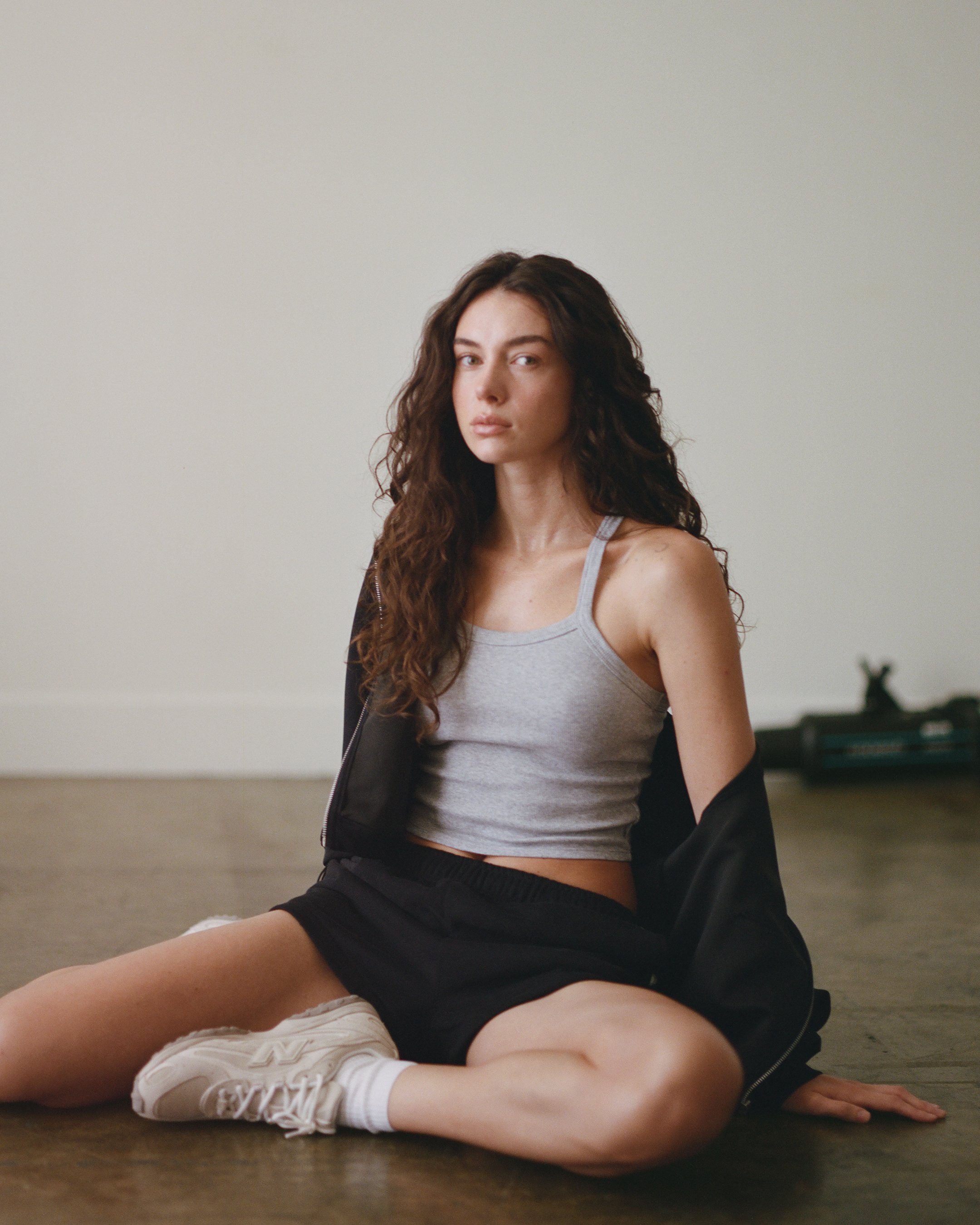 Young woman with long curly hair sitting on the floor in a minimalist room, wearing a gray tank top and black shorts, with a black jacket draped over her shoulders, looking at the camera.