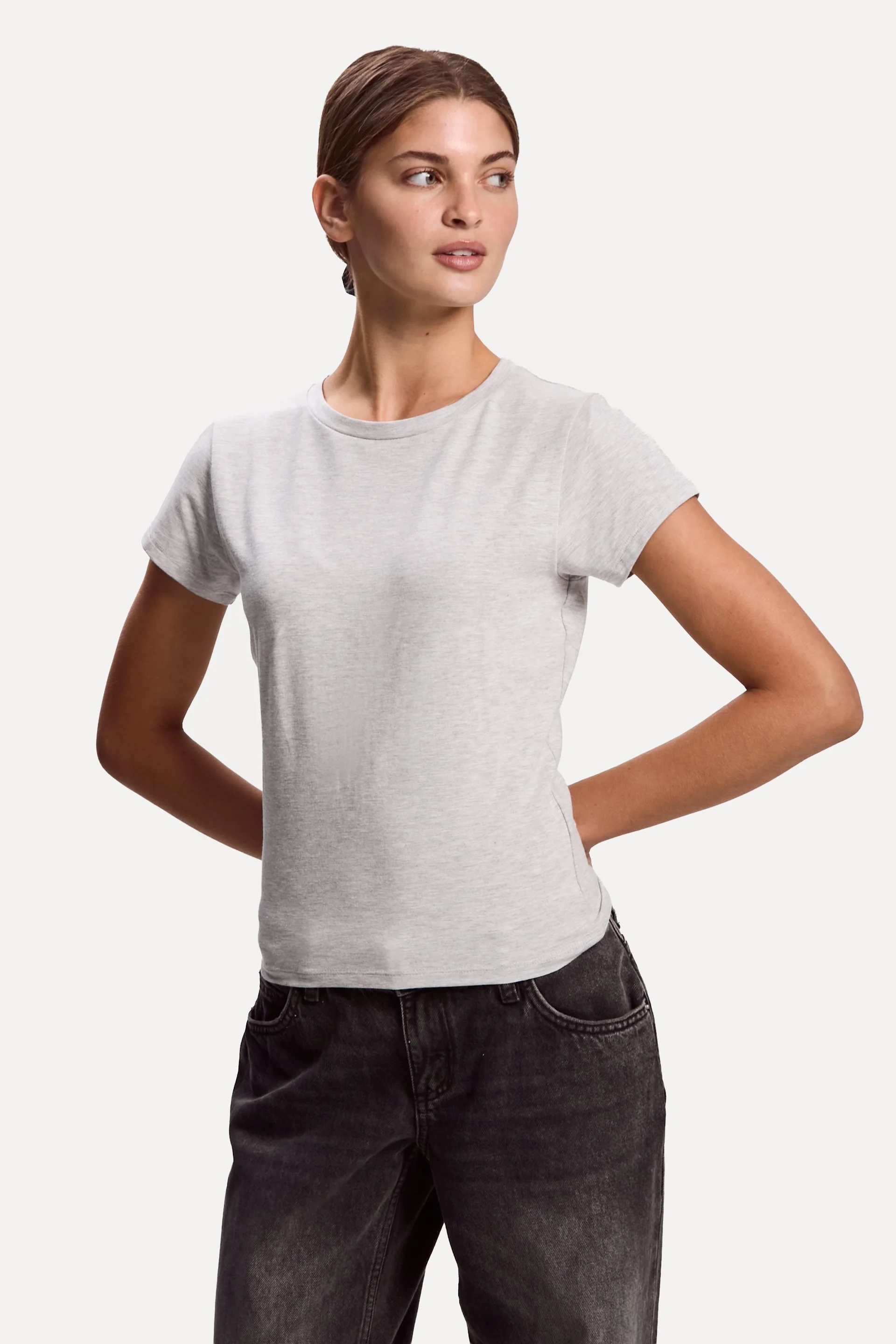 A woman with brown hair styled in a braid, wearing a light gray t-shirt and black jeans, standing with her hands behind her back against a plain white background.