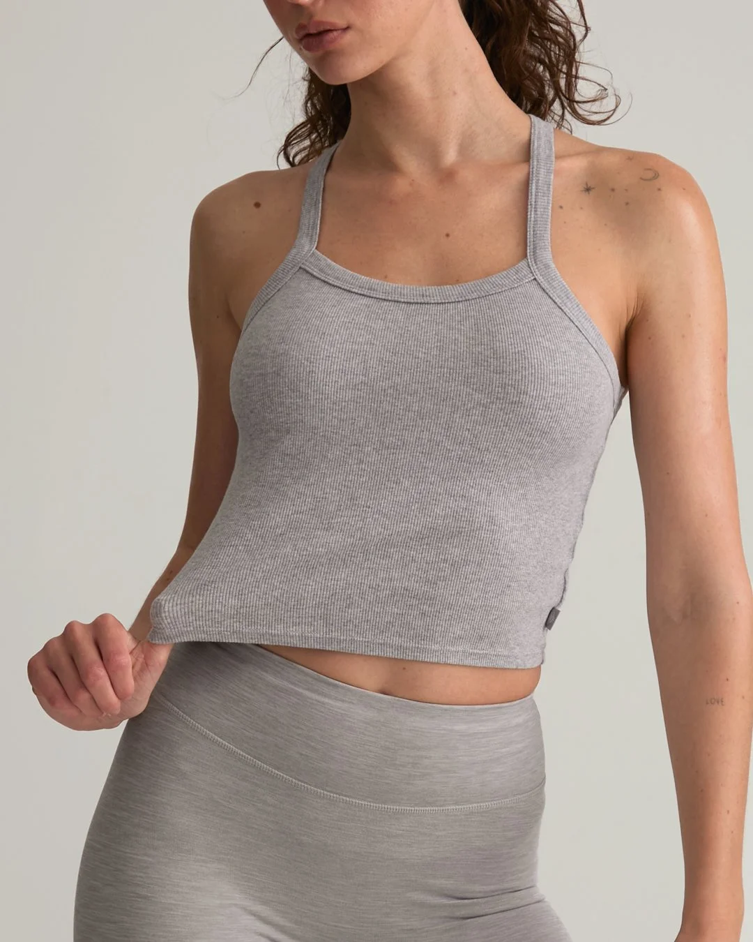 A woman wearing a light gray tank top and matching leggings, holding the hem of her tank top with her left hand, against a plain light background.