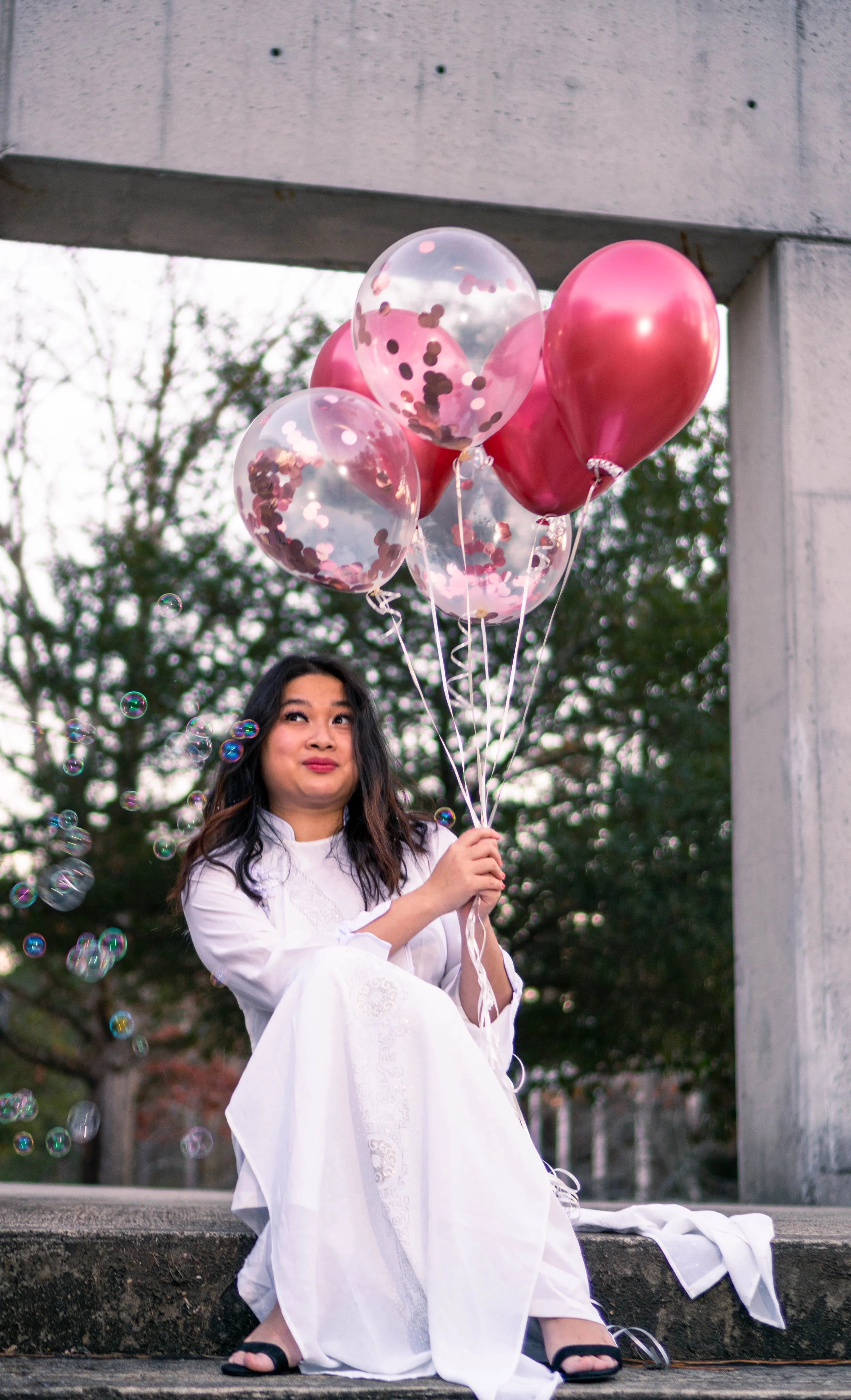 A woman in a white dress sitting on a concrete ledge holding a bunch of pink and clear balloons with confetti inside, with soap bubbles floating around, outdoors with trees in the background.