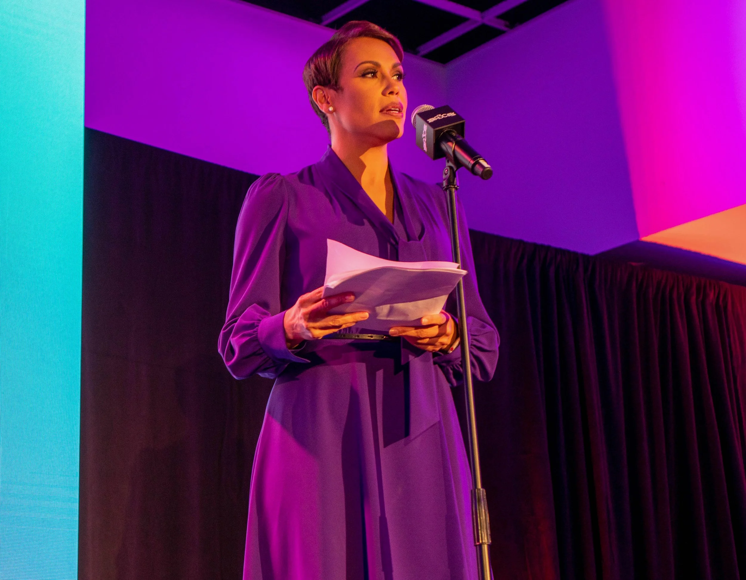 A woman in a purple dress speaking at a microphone on stage, holding a stack of papers, with purple and pink lighting.