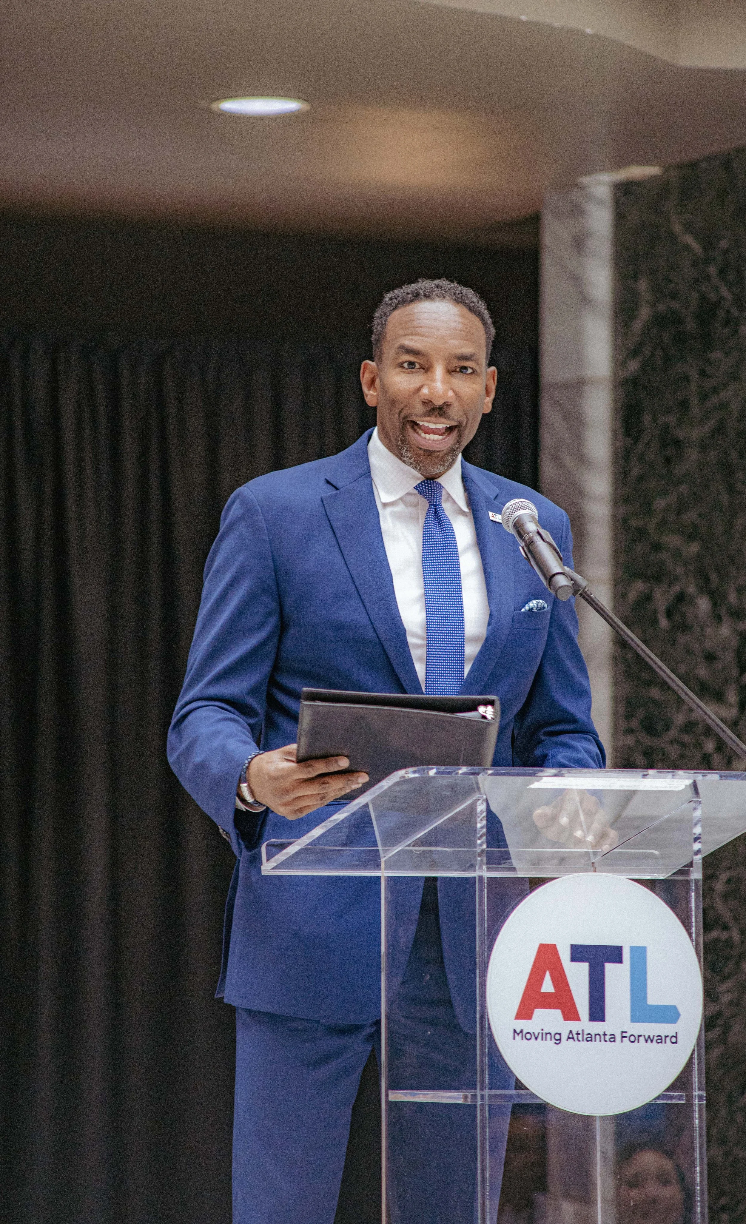 A man in a blue suit speaking at a podium with a microphone, with a logo for ATL and the words 'Moving Atlanta Forward' on the front.