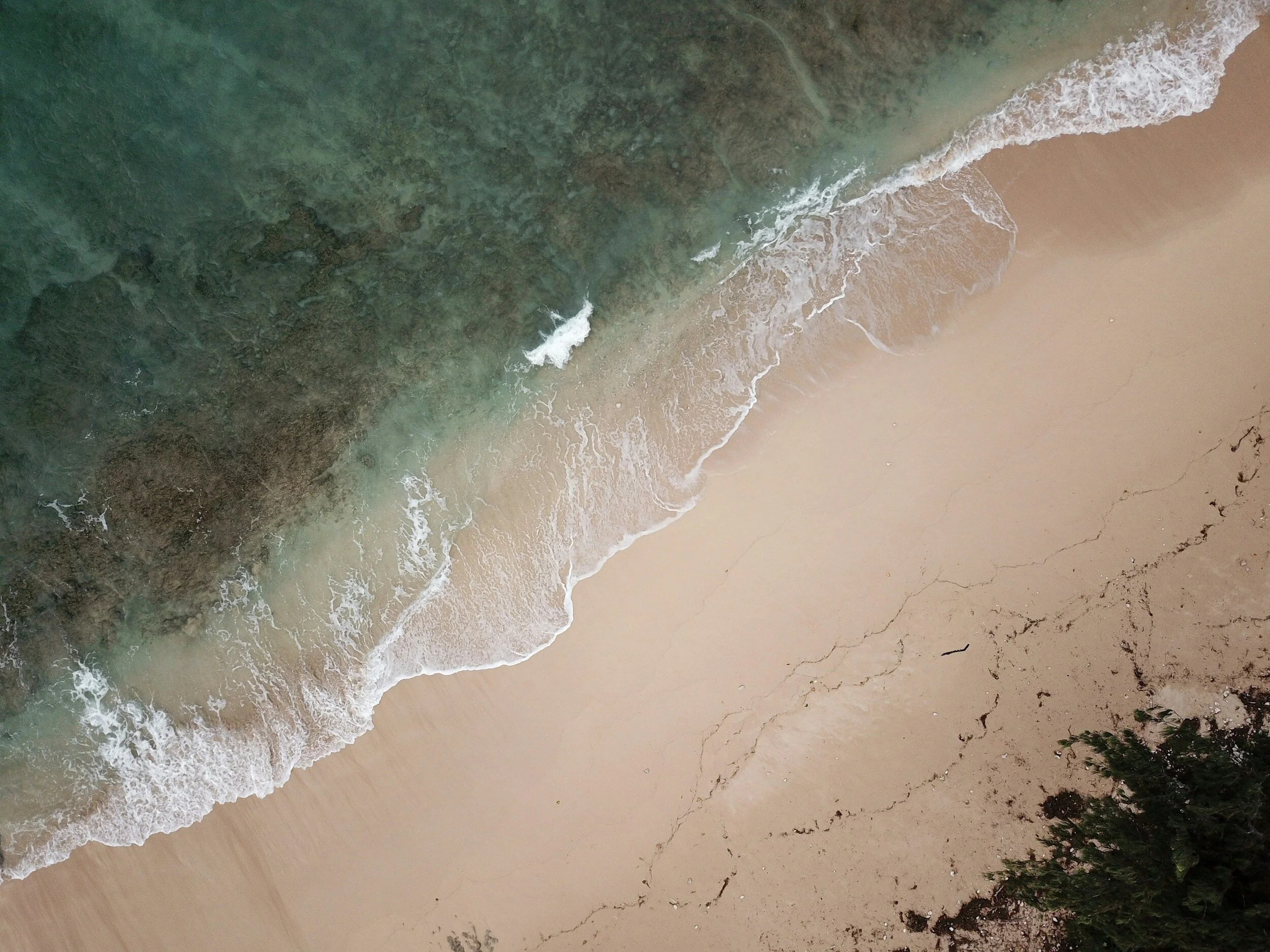 Aerial view of a sandy beach with waves crashing onto the shore and greenery in the bottom right corner.