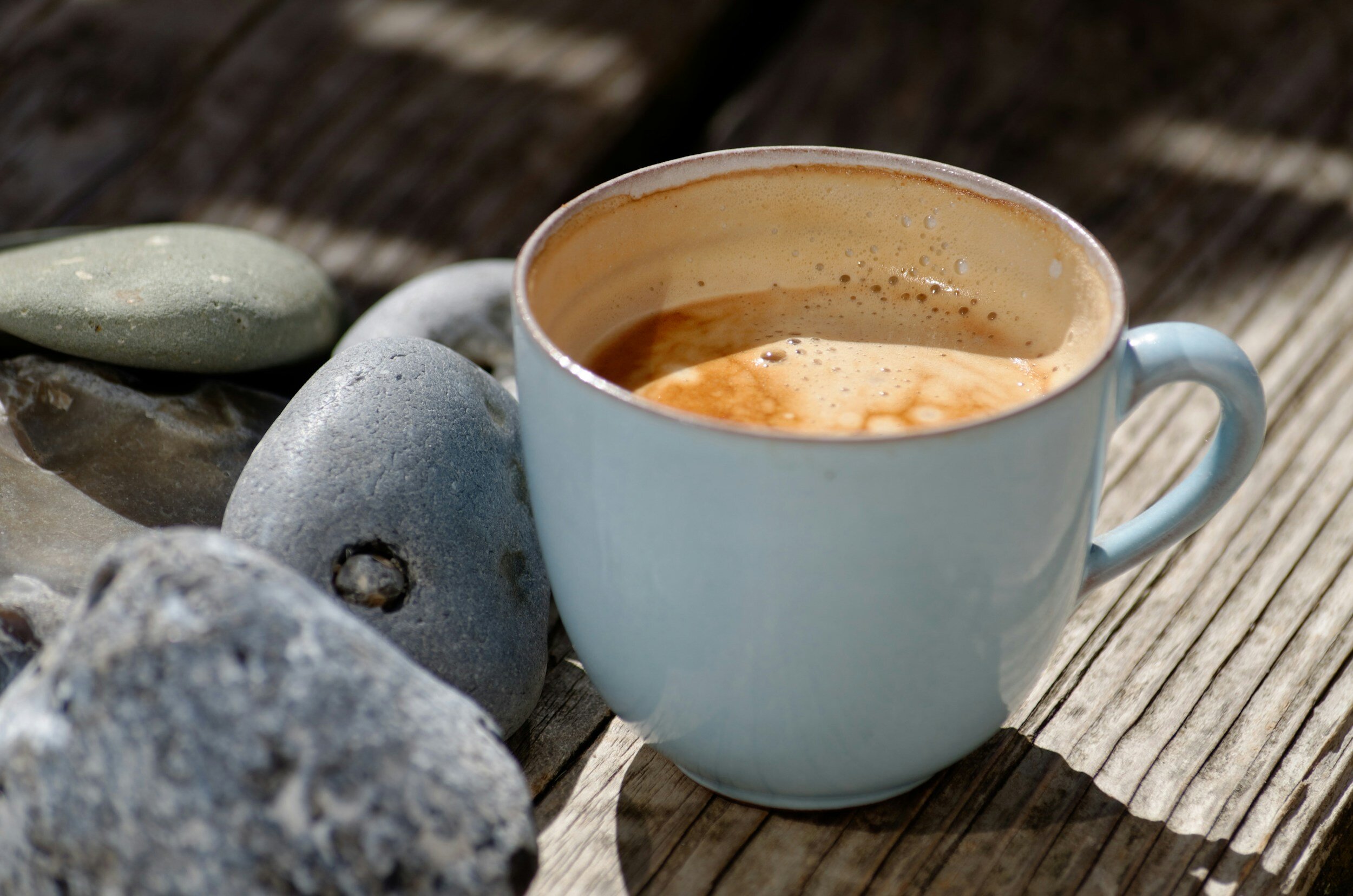 A blue ceramic mug filled with coffee, placed on a weathered wooden surface, surrounded by smooth gray and green stones.