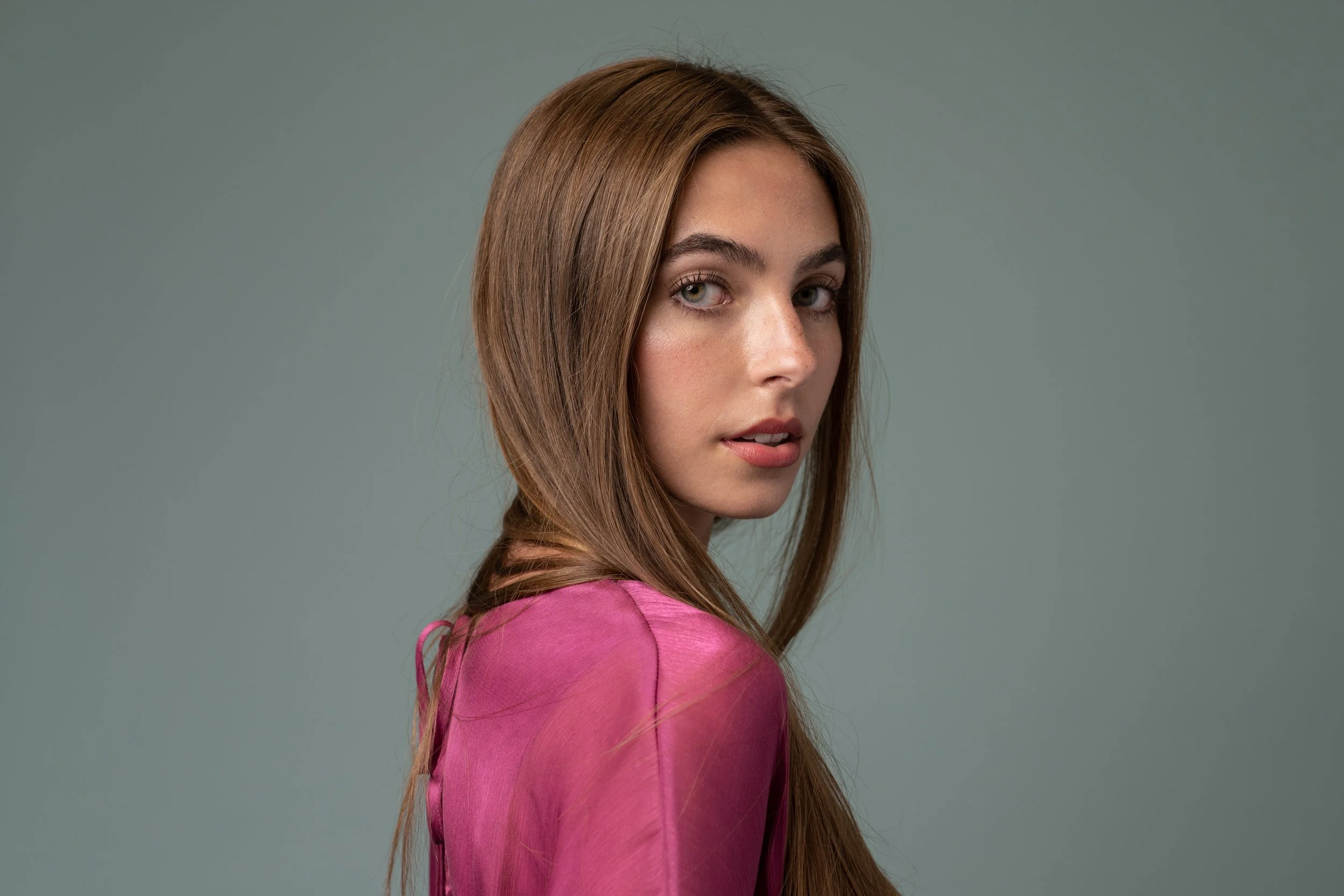 A woman with long, light brown hair and blue eyes looks over her shoulder at the camera, wearing a pink satin top against a plain grey background. Photo by Brock Byers
