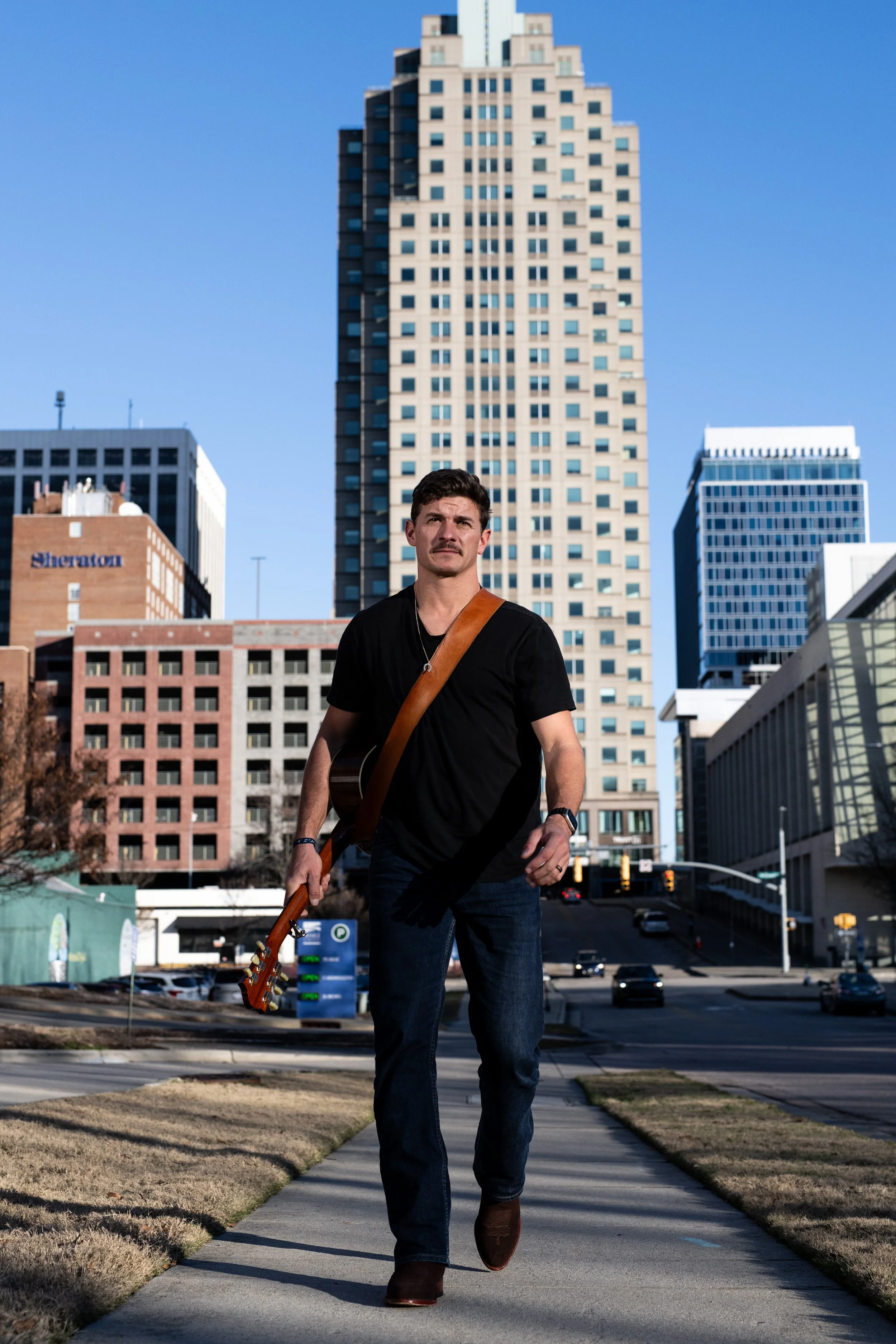 Chris Lewis, musician, walking on a city sidewalk carrying a guitar with a city skyline background. Photo by Brock Byers 