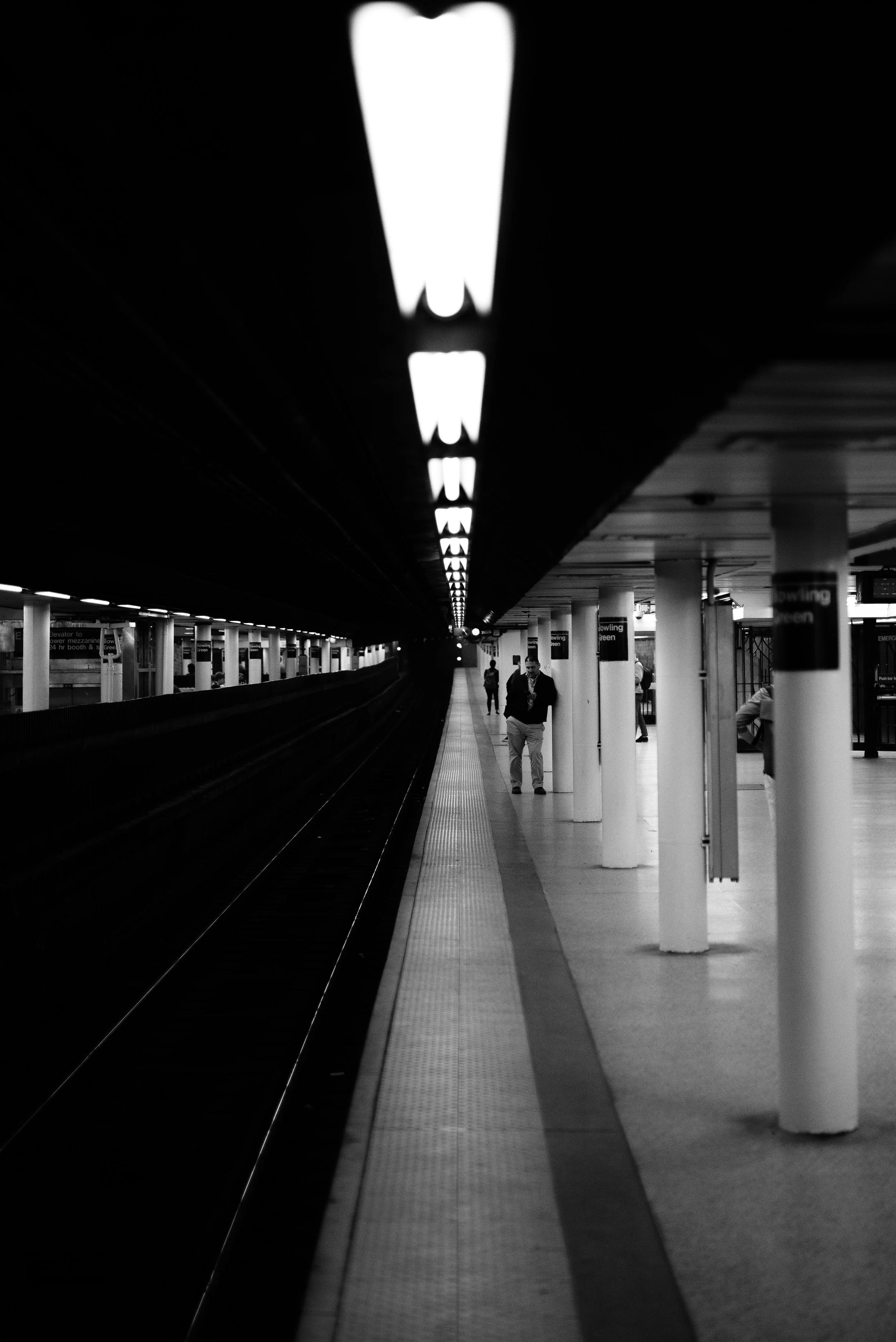 Black and white photo of a subway station platform with several people waiting. The platform has a row of bright overhead lights and white columns supporting the ceiling. The train tracks are visible on the left side, fading into the distance. Photo 