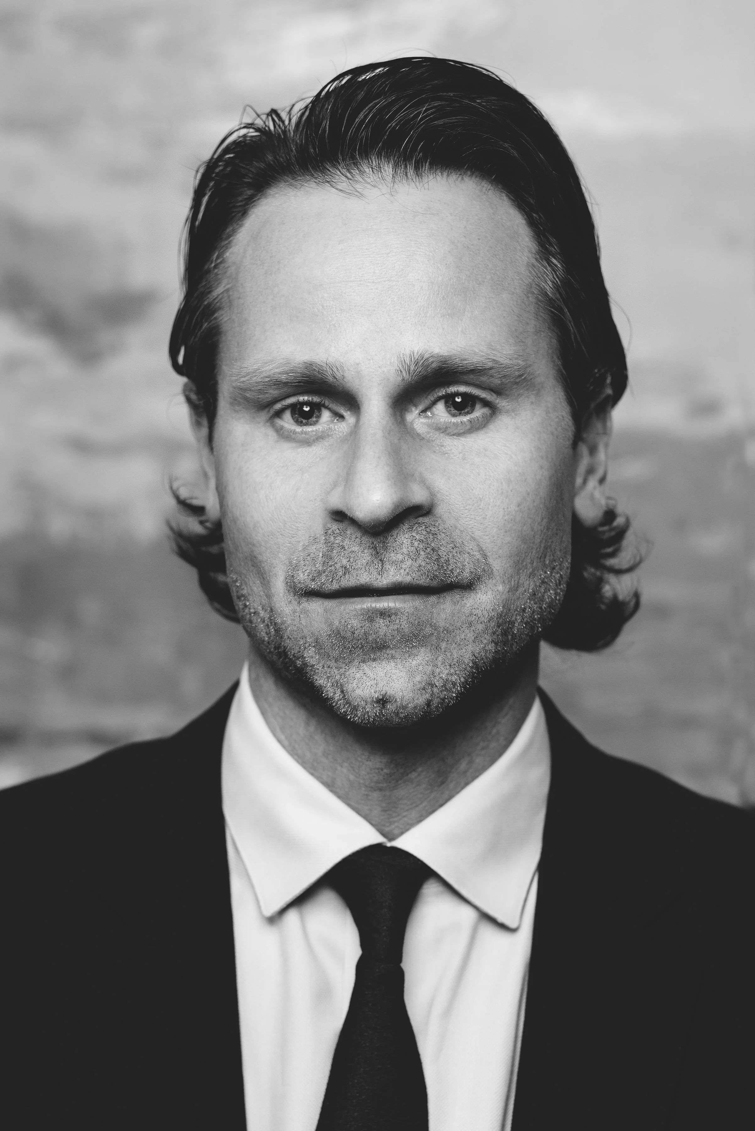 Black and white portrait of a man with dark, wavy hair, wearing a suit, white shirt, and tie, looking directly at the camera. Photo by Brock Byers 