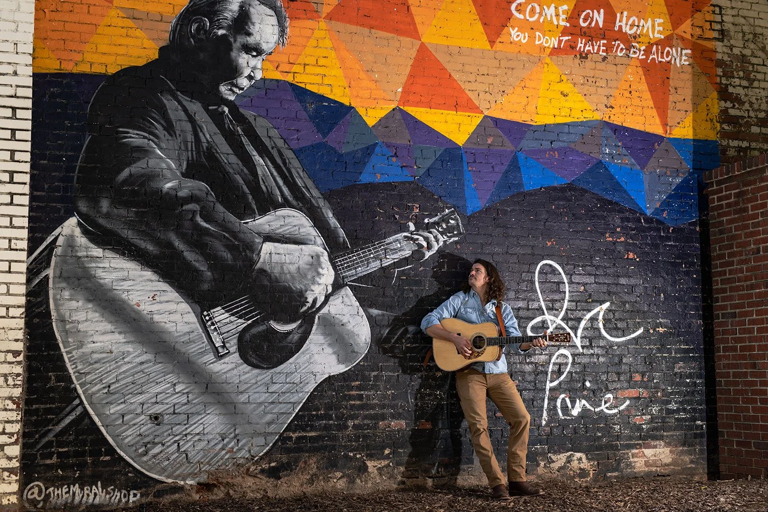 A musician with brown curly hair, wearing a light blue shirt and brown pants, playing an acoustic guitar while standing against a large mural on a brick wall. The mural features a black and white portrait of a man playing an acoustic guitar, with a c
